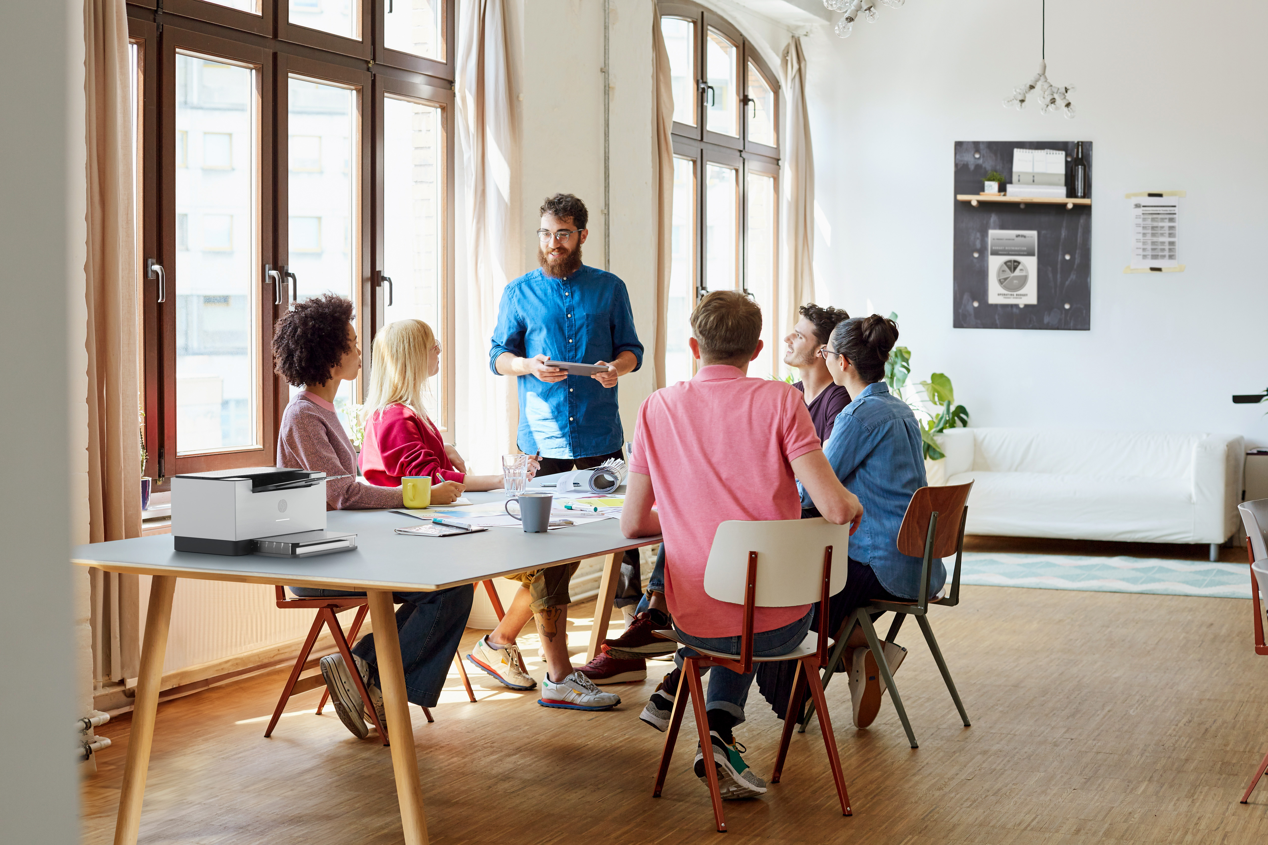 Een vergadering is aan de gang. Een man met een baard presenteert. Anderen zitten aan een tafel, sommigen maken aantekeningen.