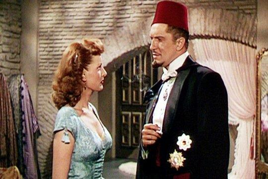 Ein Mann in Fez und Smoking steht einer Frau in blauem Kleid gegenüber. Sie befinden sich in einem Raum mit Backsteinmauer und einer dekorativen Tür.