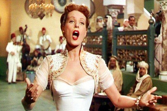 Eine Frau in weißem Kleid singt. Eine Band spielt hinter ihr. Menschen in traditioneller Kleidung sind im Hintergrund.