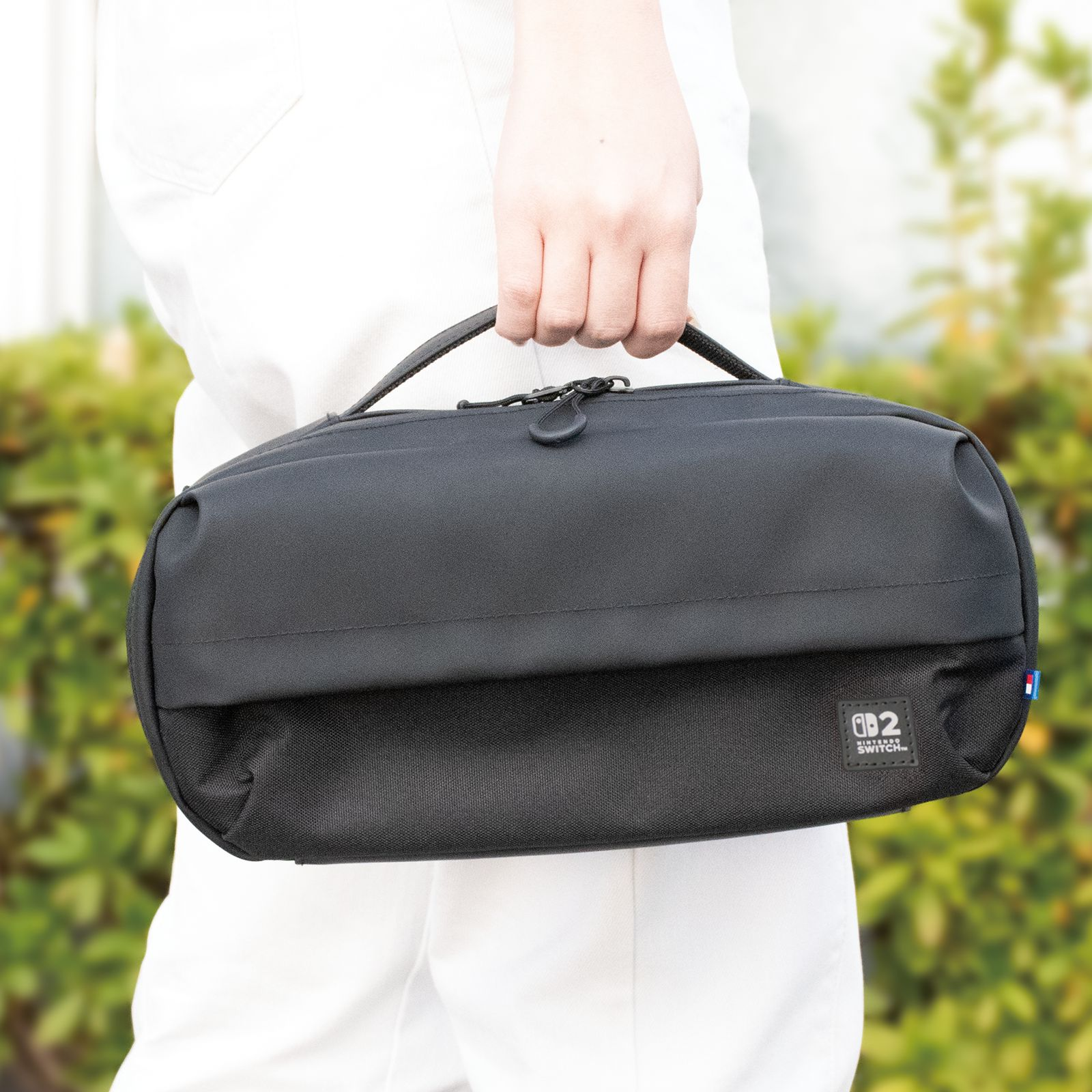 Eine Person hält eine schwarze Nintendo Switch-Tragetasche. Weiße Hose ist sichtbar. Grünes Laub bildet den Hintergrund.
