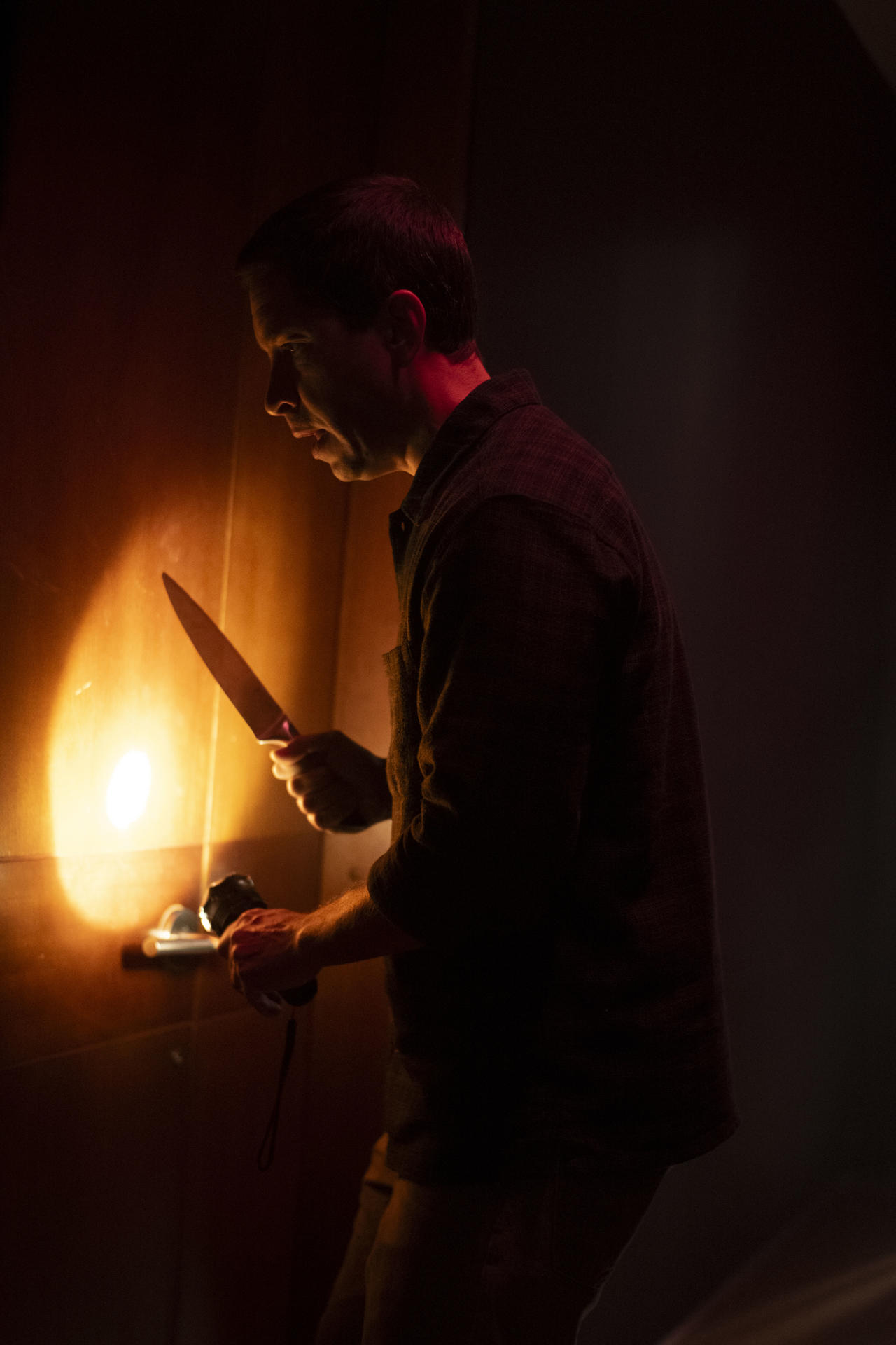 Ein Mann hält ein Messer und eine Taschenlampe, beleuchtet von einer Lichtquelle. Er befindet sich in einem schwach beleuchteten Raum, wahrscheinlich in einer spannungsgeladenen Situation.