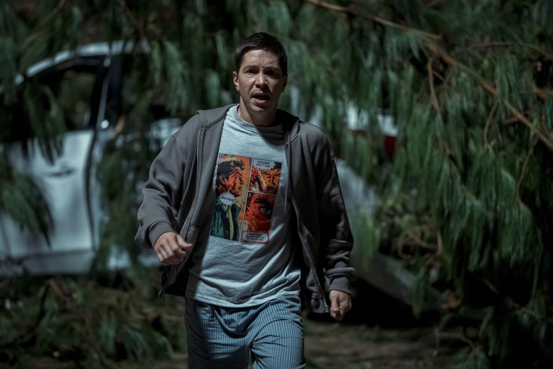 Mann in grauem Hoodie und T-Shirt rennt auf die Kamera zu. Hinter ihm sind ein Auto und Grün. Das T-Shirt hat einen Comic-Strip-Druck.
