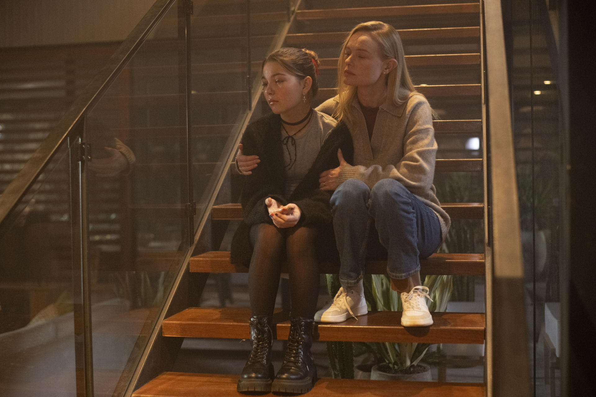 Zwei Frauen sitzen auf einer Treppe. Eine tröstet die andere. Holztreppe, Glasgeländer und neutrale Töne dominieren.