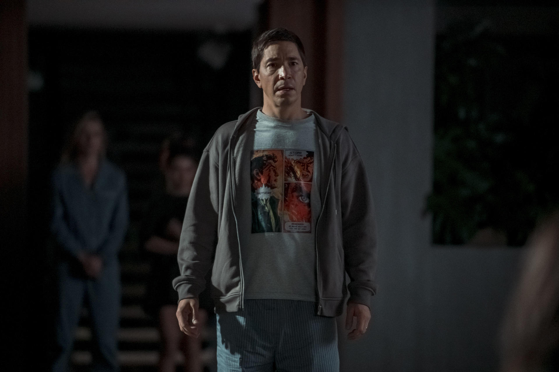 Ein Mann mit grauem Hoodie und Comic-Shirt steht draußen. Zwei verschwommene Figuren stehen hinter ihm. Die Szene ist dunkel.