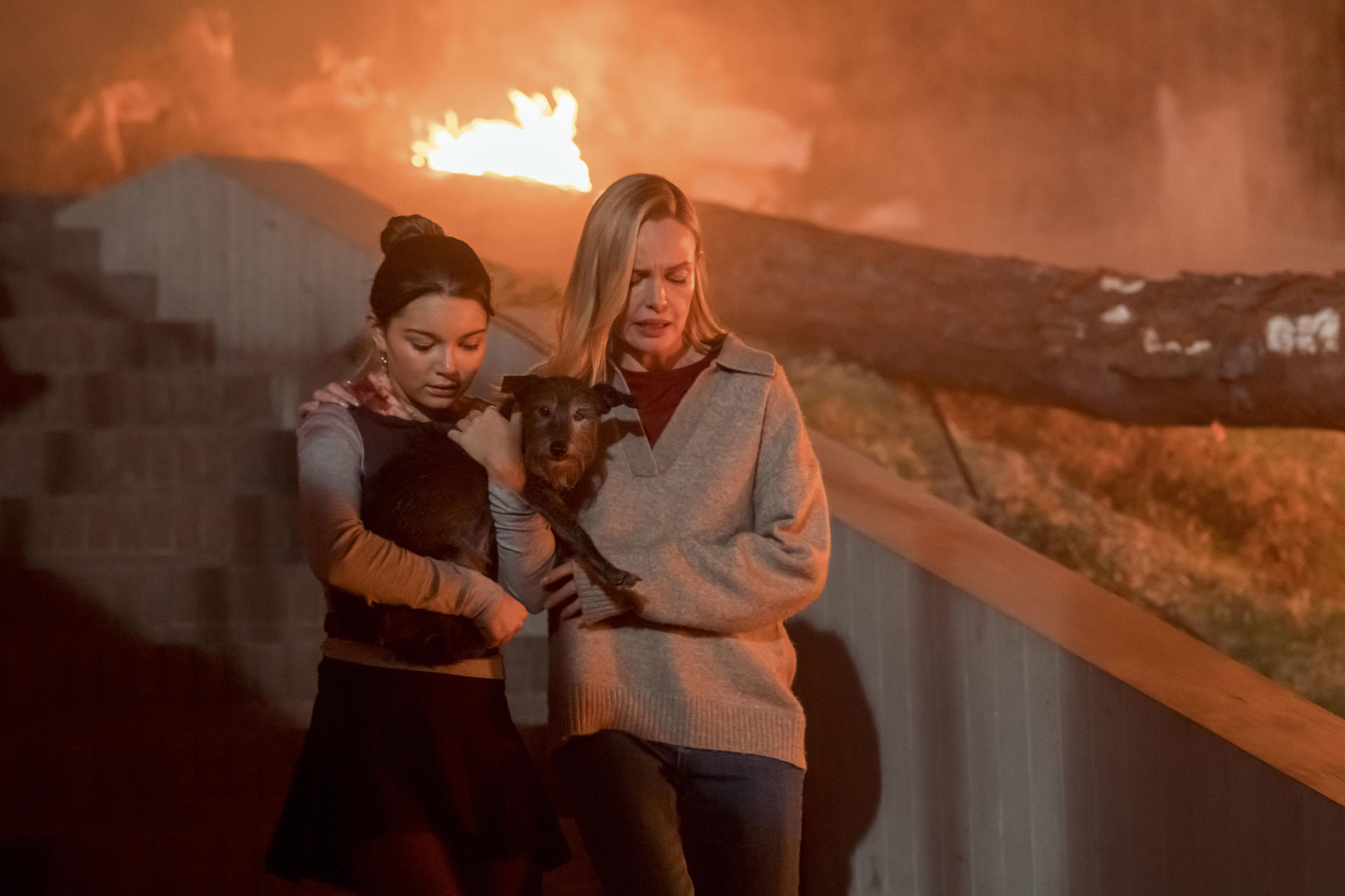 Zwei Frauen halten einen Hund, im Hintergrund ein Feuer. Eine Frau ist blond, die andere hat dunkles Haar.