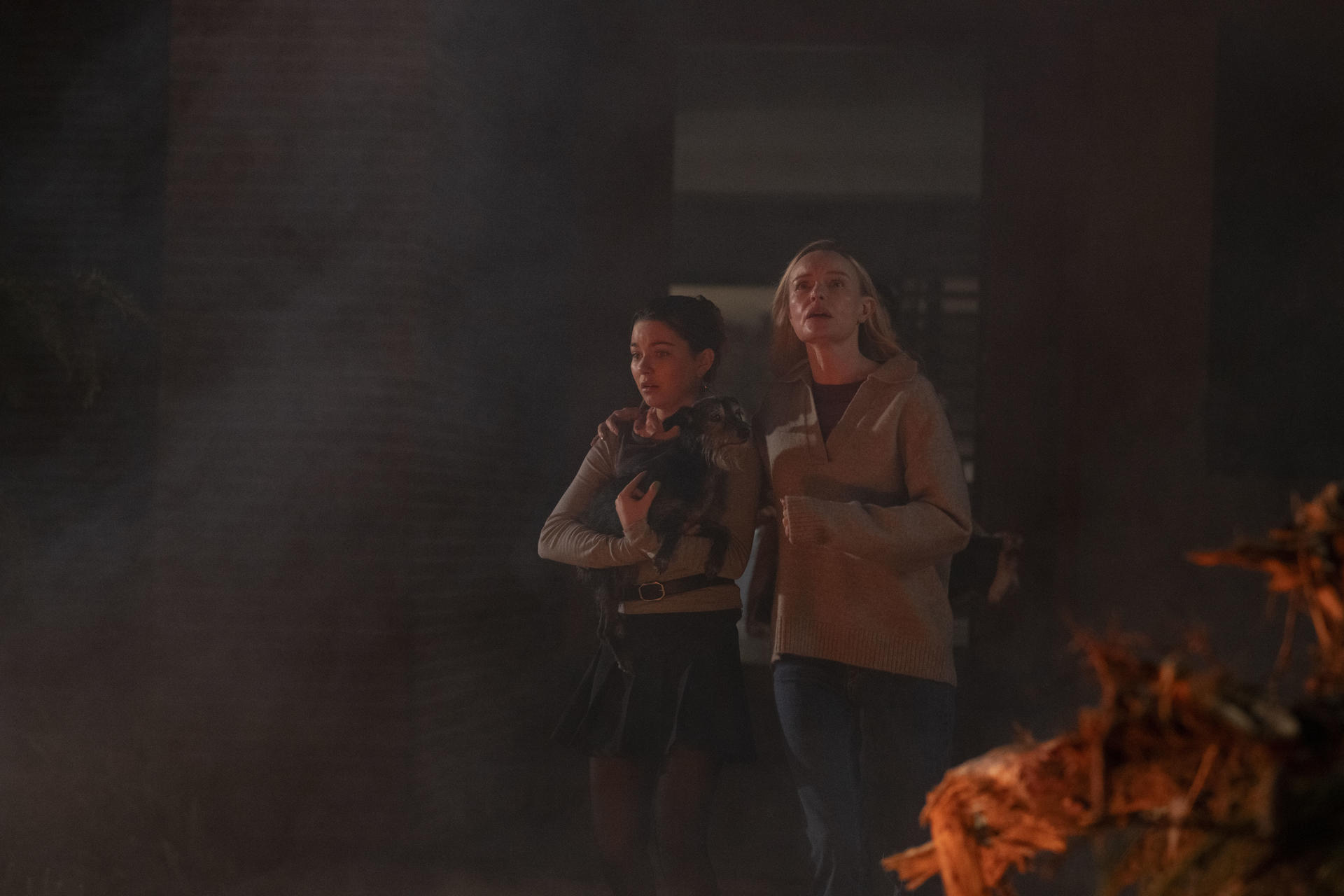 Zwei Frauen, eine mit Hund, sehen besorgt aus. Sie stehen in der Nähe eines Feuers. Eine trägt einen Pullover und Jeans, die andere einen Rock.