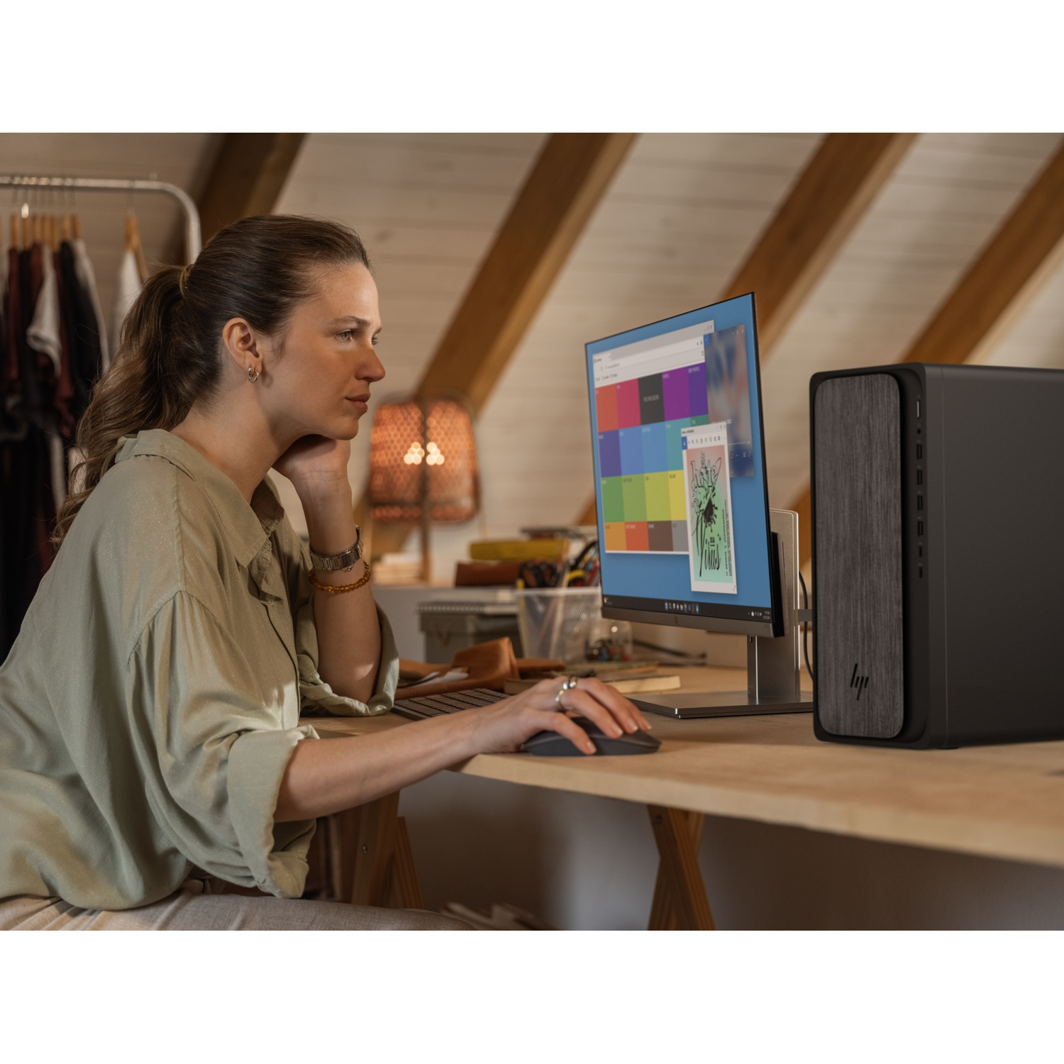 Vrouw achter bureau met computer. Monitor toont kleurrijke iconen. Zwarte pc-toren rechts. Houten bureau met kledingrek op de achtergrond.
