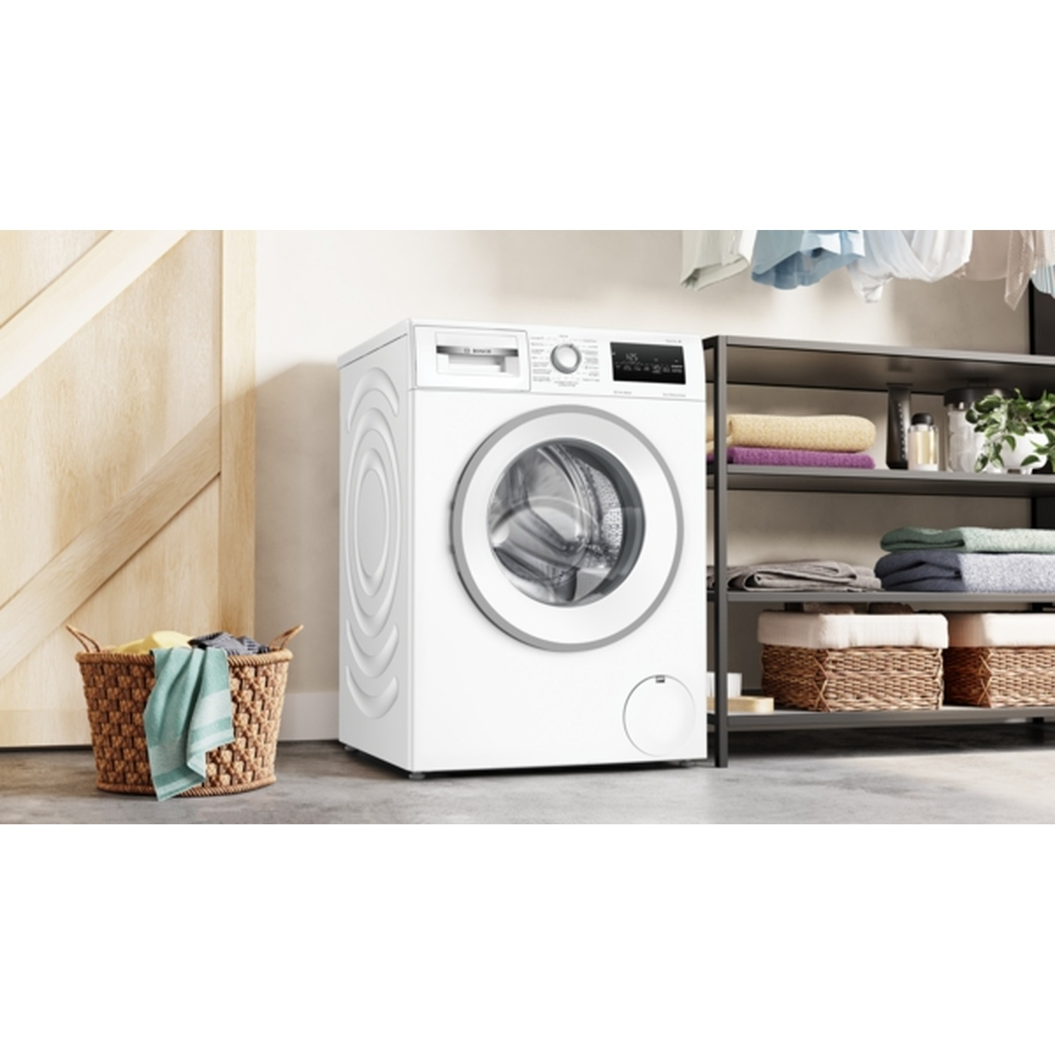 A white washing machine stands next to a wooden door and a metal shelf with towels and baskets. A basket with clothes sits on the floor.