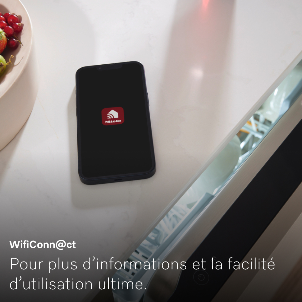 Un smartphone affiche l'application Miele sur une surface blanche à côté d'un réfrigérateur. Un bol de fruits à gauche.