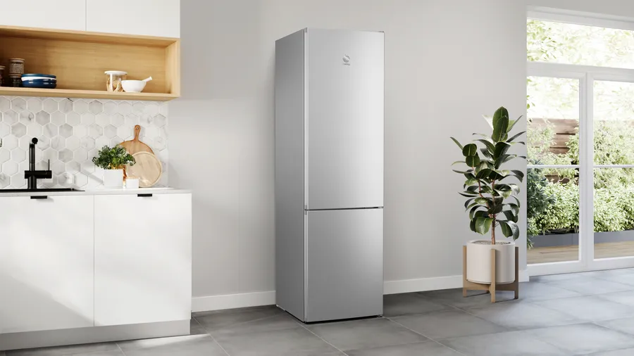 Cocina moderna con refrigerador plateado, planta y ventana al jardín. Gabinetes blancos y estantes de madera.