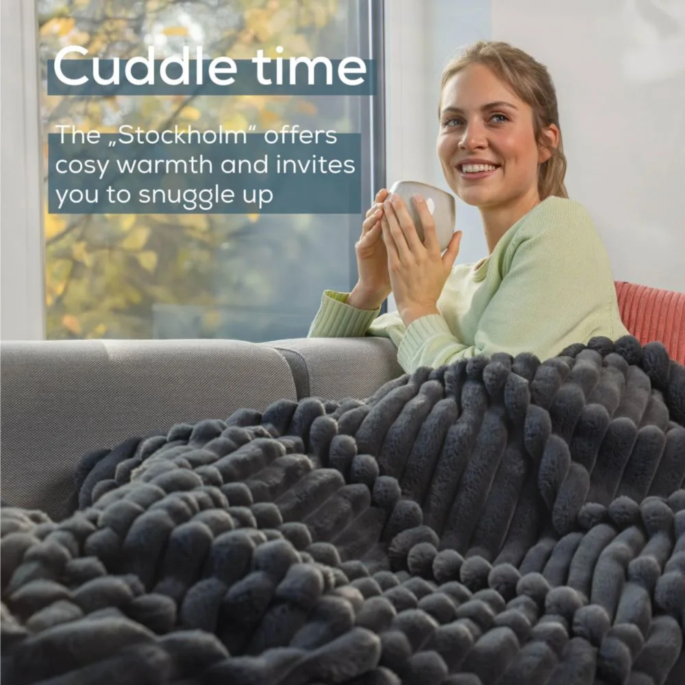 Donna sorridente, tazza in mano, avvolta in coperta grigia, seduta su divano. Testo: Tempo di coccole, offre calore.