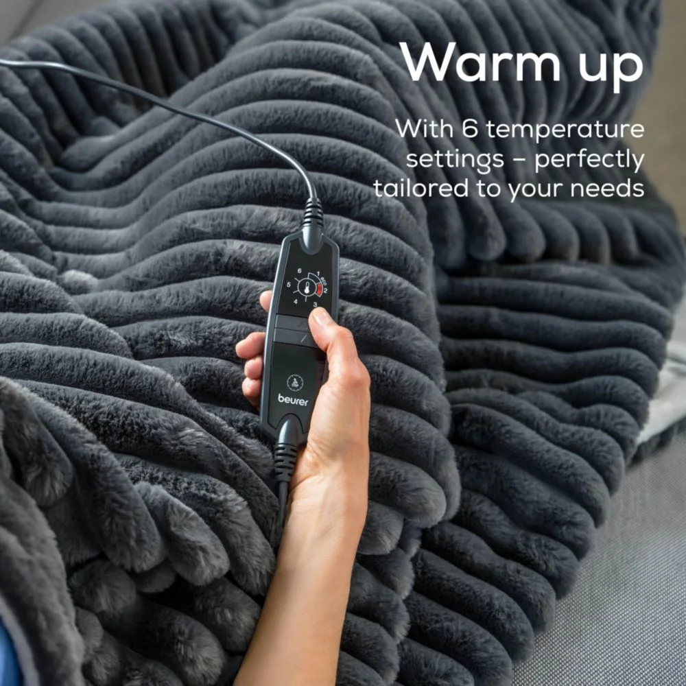 Una mano tiene in mano il controller di una coperta riscaldata grigia. Il controller mostra le impostazioni della temperatura. La coperta ha una trama ondulata.