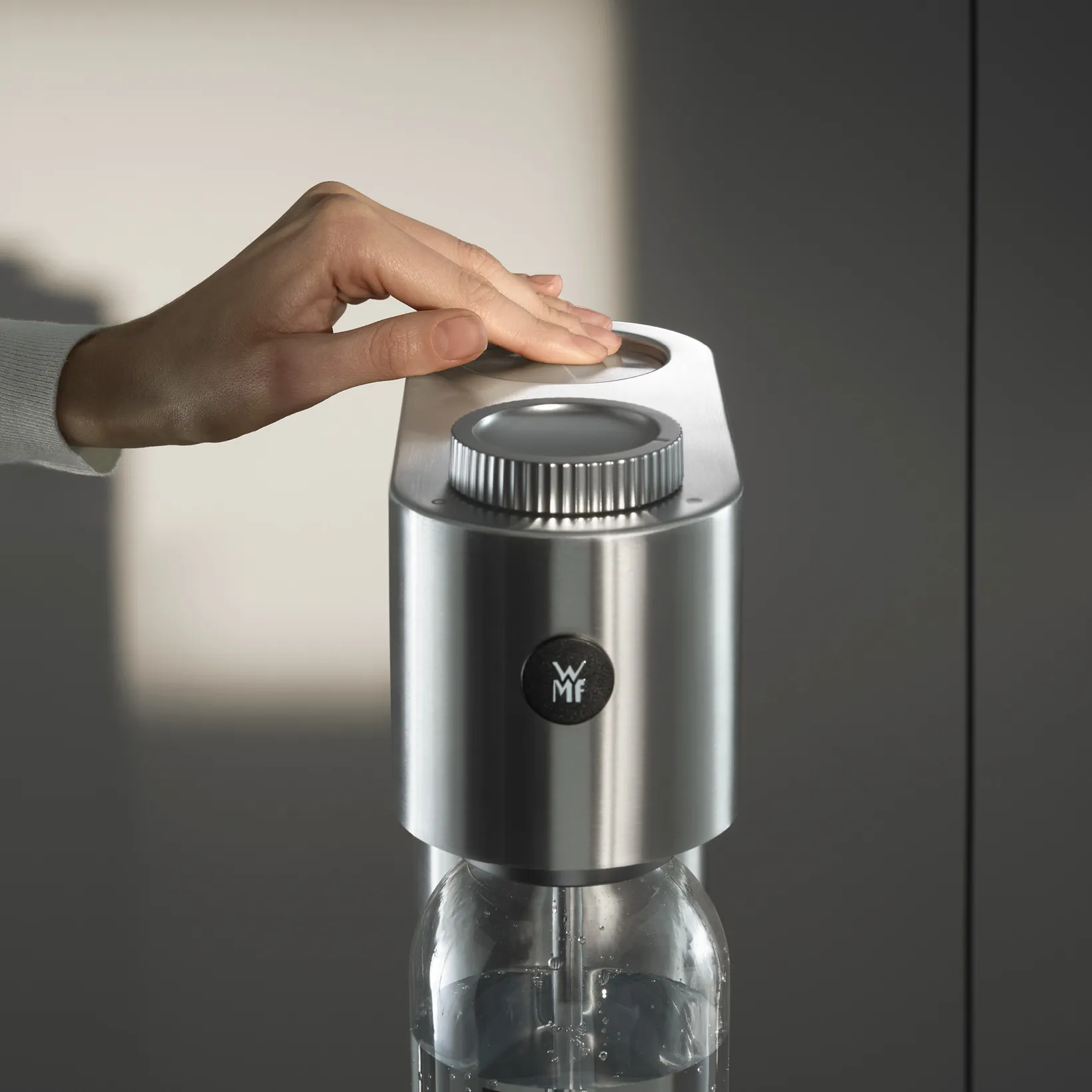 Une main appuie sur le dessus argenté d'une machine à soda. La machine a un bouton et une bouteille transparente. Fond gris.