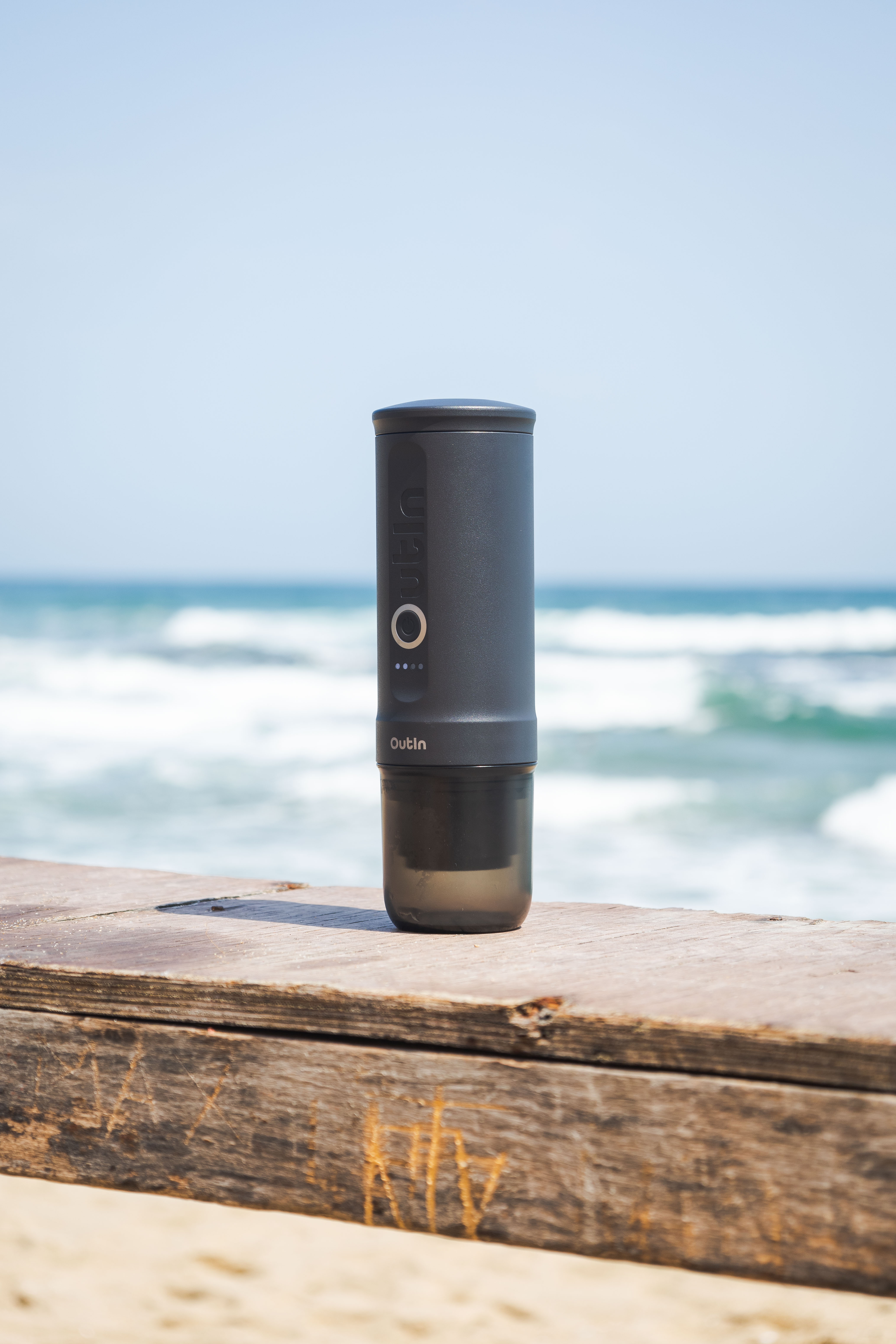 Ein schwarzer Outin-Kaffeezubereiter steht auf einer Holzplanke am Strand. Ozean und Himmel sind im Hintergrund.