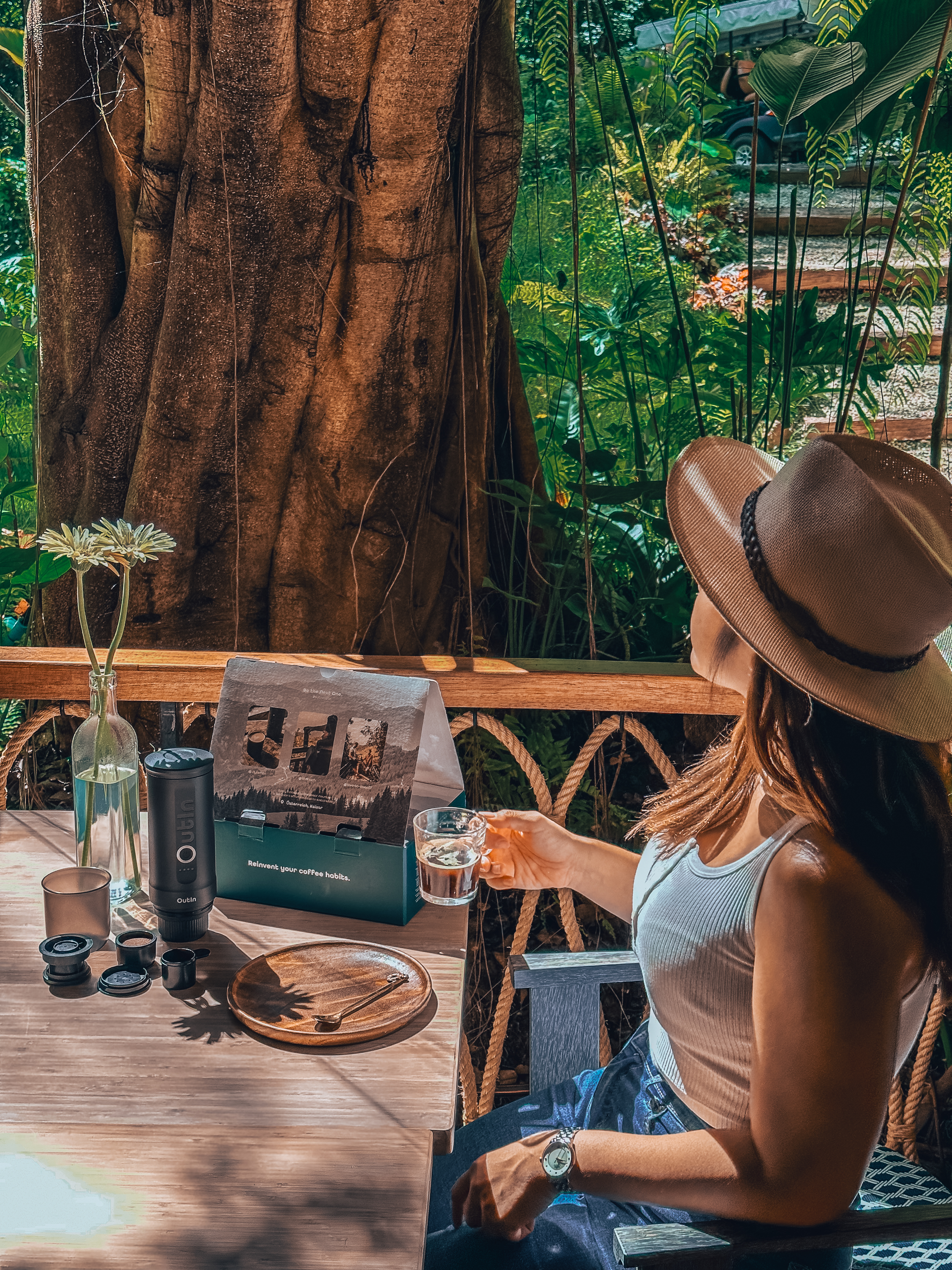 Eine Frau mit Hut hält Kaffee und sitzt an einem Holztisch mit Kaffeemaschinenausrüstung. Im Hintergrund ein Baum und üppiges Grün.