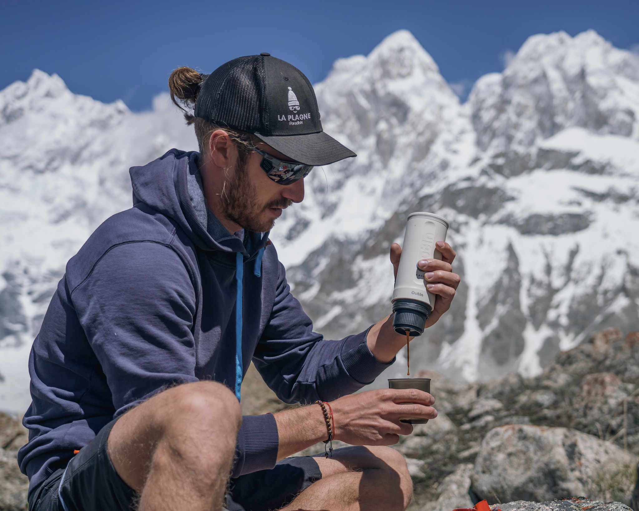 Mann in Kappe gießt Kaffee in eine Tasse, schneebedeckte Berge im Hintergrund. Klarer blauer Himmel.
