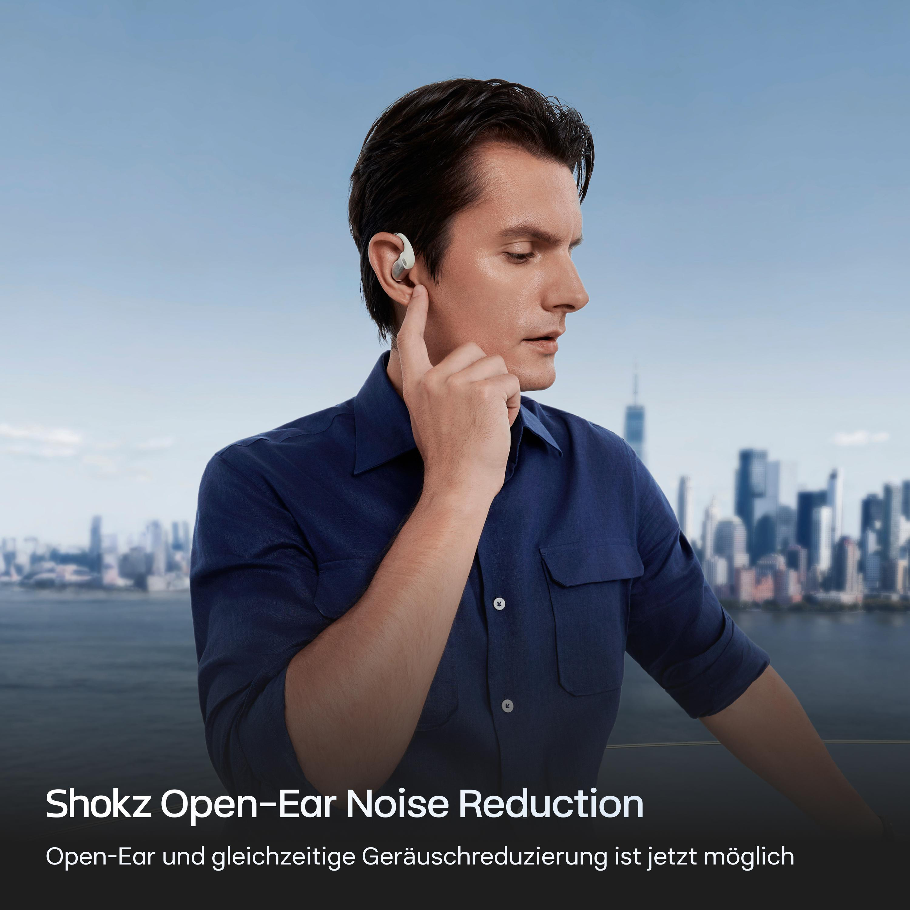 Ein Mann mit Kopfhörern berührt sein Ohr. Im Hintergrund ist eine Skyline zu sehen. Text: Shokz Open-Ear Noise Reduction.