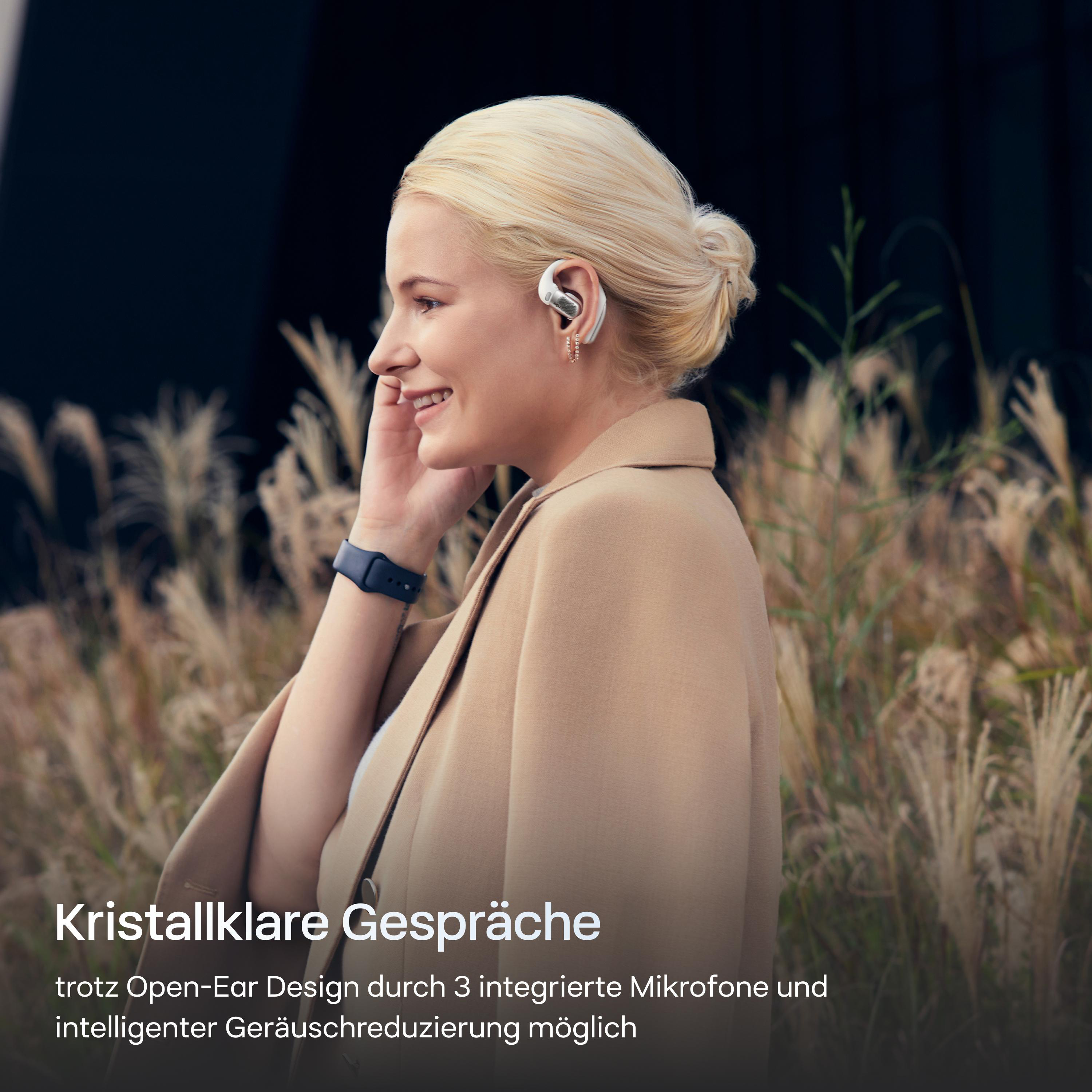 Blonde Frau mit Kopfhörern, lächelnd. Trägt einen beigen Blazer. Hohes Gras im Hintergrund.