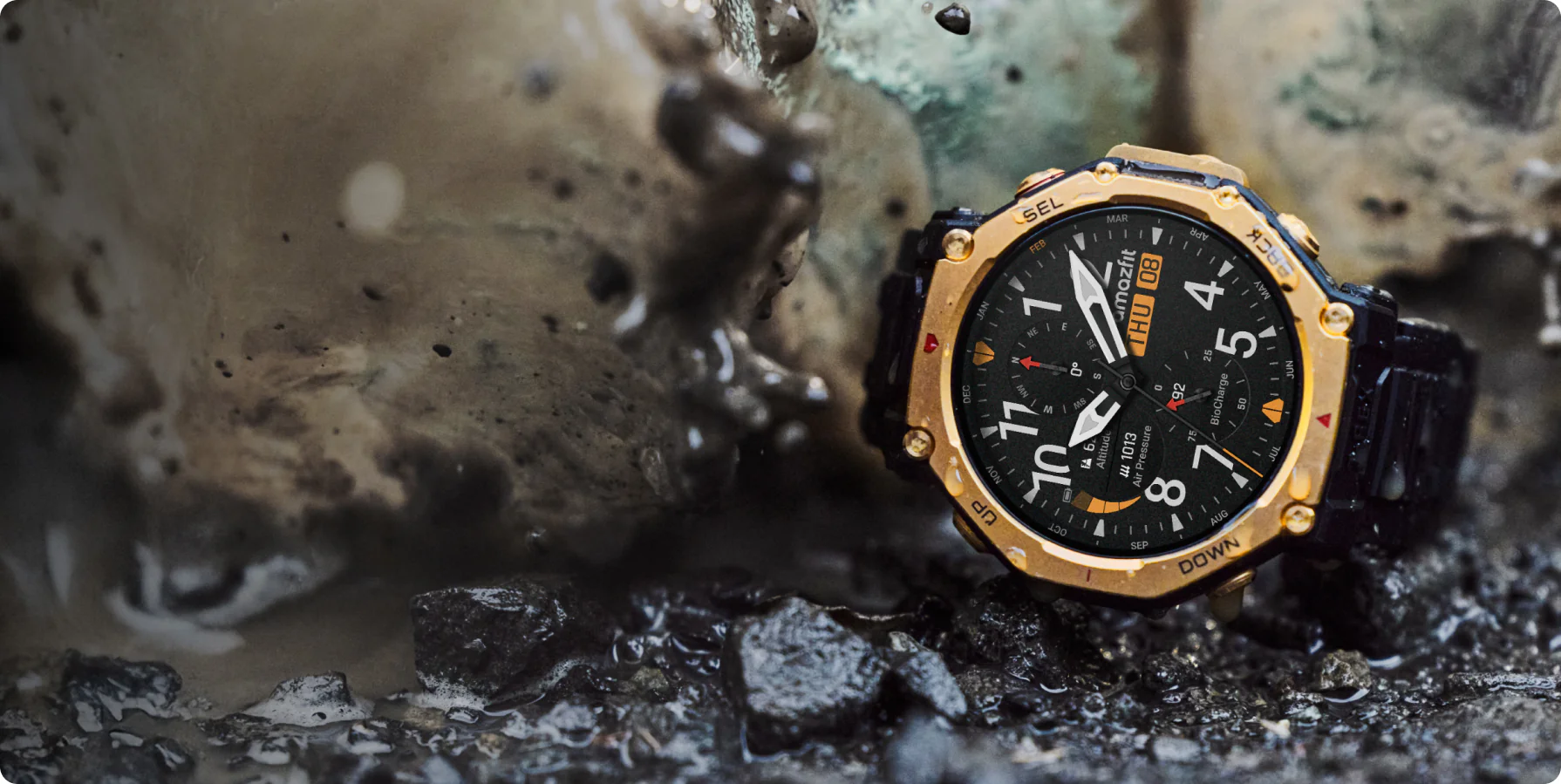 Une smartwatch Amazfit dorée et noire est posée dans la boue et des petites roches. La montre affiche l'heure et des données, avec des gouttes d'eau.