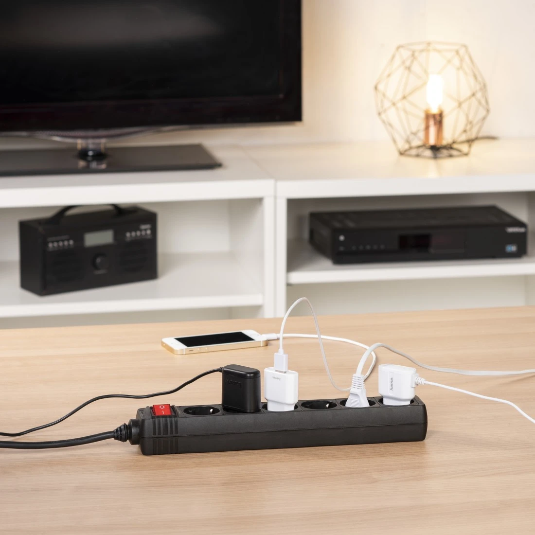 Una regleta con varios enchufes está sobre una mesa de madera. Tiene cargadores y un teléfono conectado. Un televisor y otros aparatos electrónicos están al fondo.