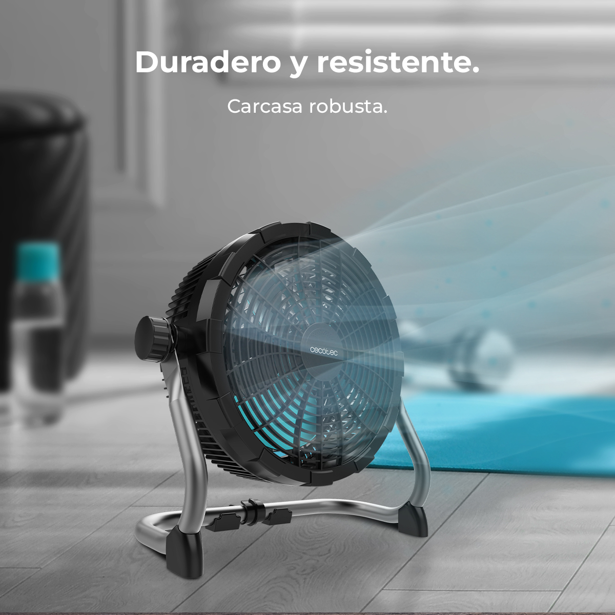 Un ventilador negro con patas plateadas está en un piso de madera. El ventilador sopla aire. Una botella de agua y una alfombra azul están en el fondo.