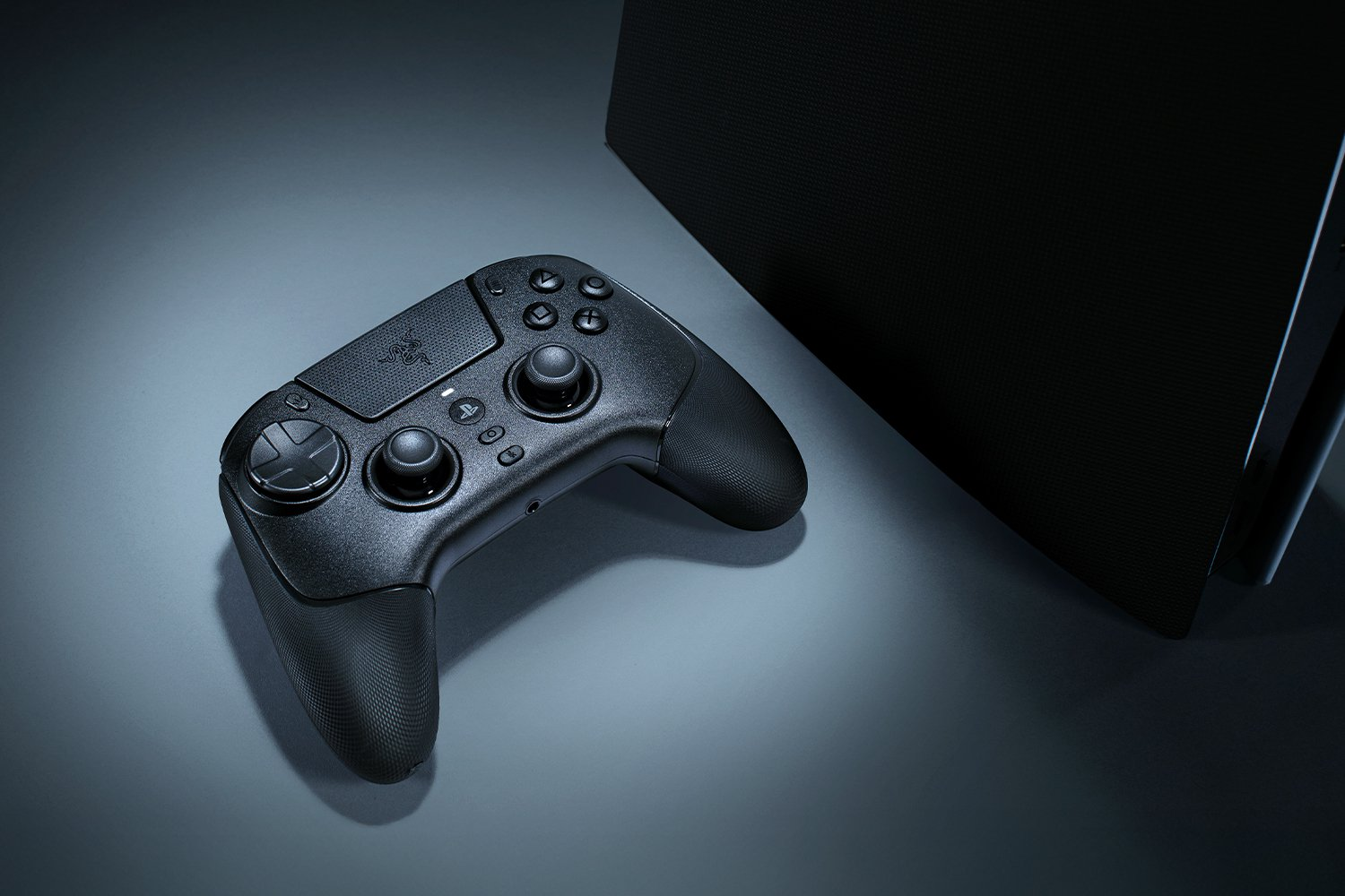 Mando Razer negro sobre una superficie gris, junto a un objeto rectangular negro. El mando tiene botones y joysticks.
