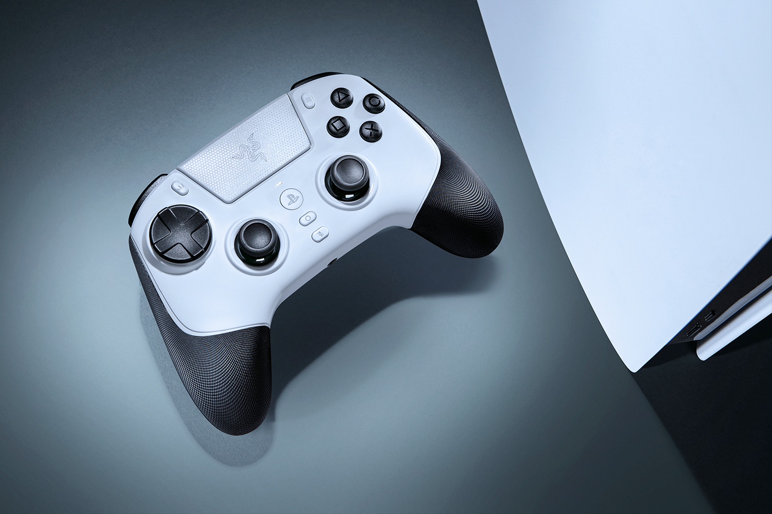 Un mando Razer blanco y negro sobre una superficie gris, con una consola de juegos blanca parcialmente visible al fondo.