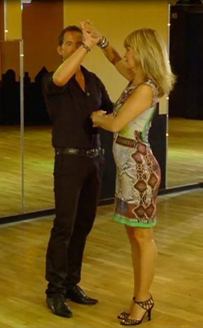 Ein Mann und eine Frau tanzen. Die Frau trägt ein Kleid und Absätze. Der Mann trägt ein schwarzes Hemd und eine Hose. Sie sind in einem Tanzstudio.