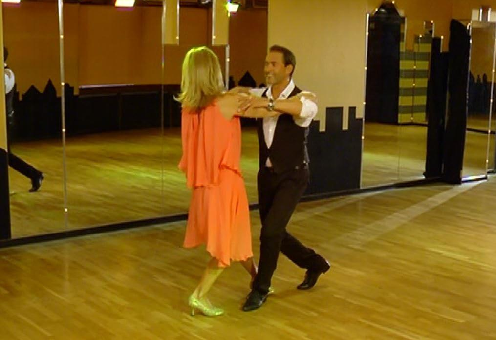 Ein Mann und eine Frau tanzen in einem Studio mit Spiegeln. Die Frau trägt ein orangefarbenes Kleid, der Mann eine Weste und schwarze Hosen.