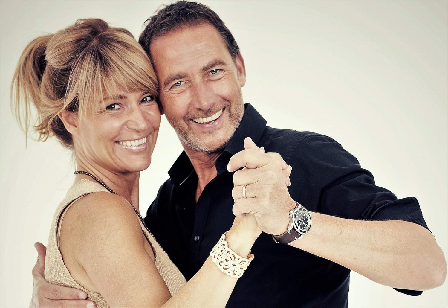 Ein lächelndes Paar tanzt. Der Mann trägt ein schwarzes Hemd und eine Uhr. Die Frau hat blondes Haar und ein Armband.
