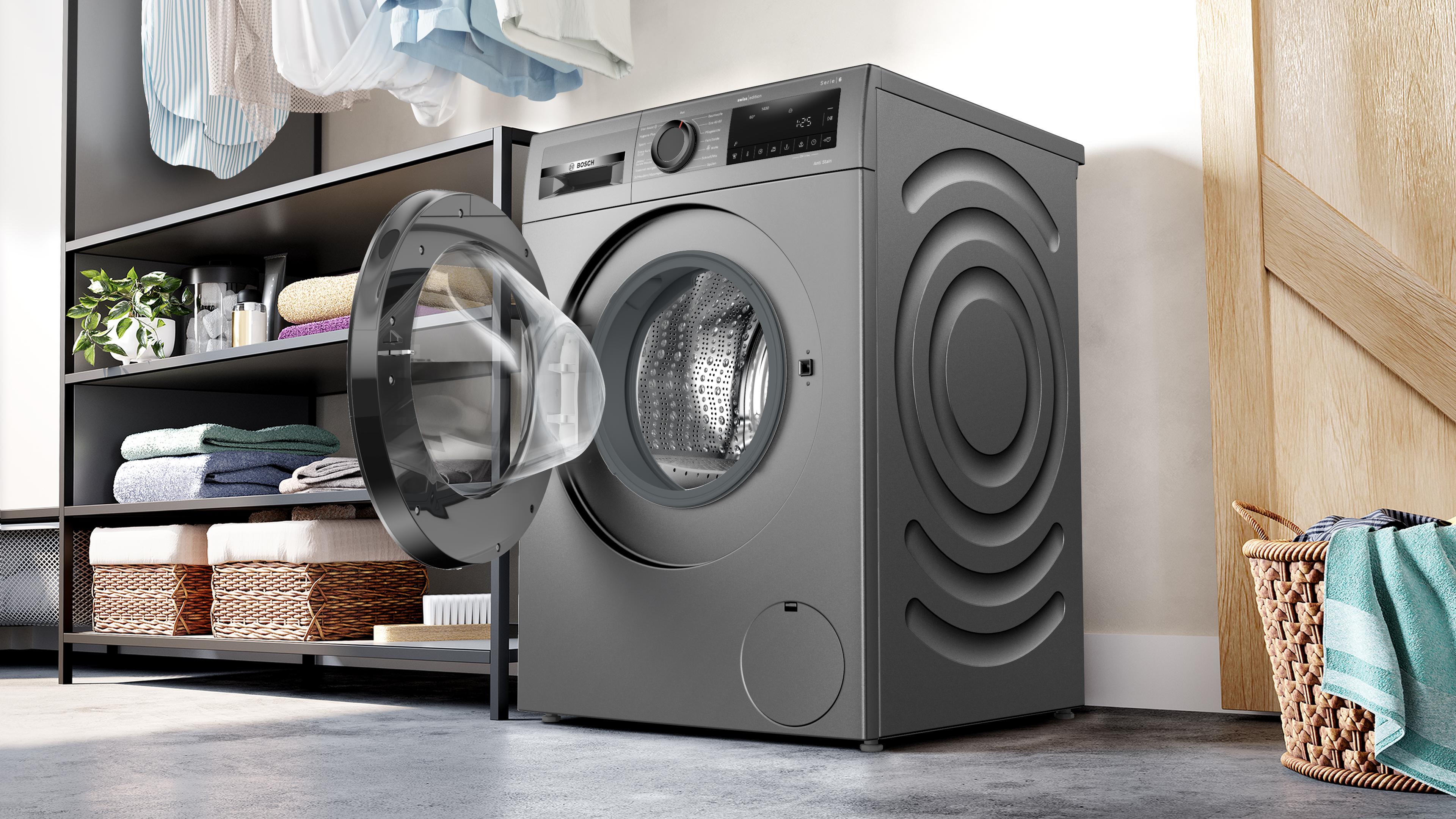 Une machine à laver grise avec une porte ouverte. Étagères avec serviettes et plantes. Vêtements suspendus. Un panier de linge.
