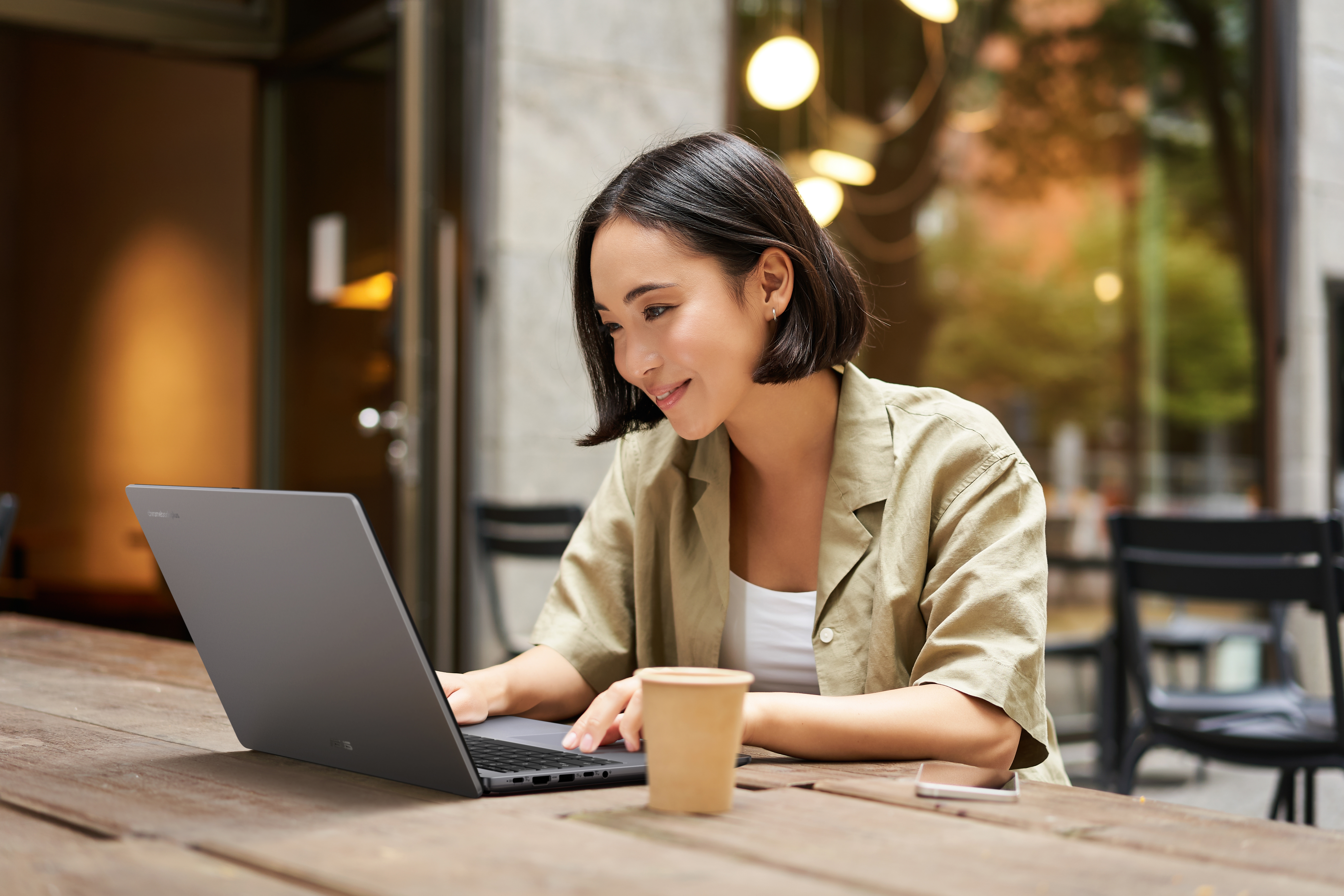 Eine Frau benutzt einen Laptop in einem Café. Sie lächelt, mit einer Kaffeetasse und einem Telefon auf dem Tisch. Draußen, verschwommener Hintergrund.