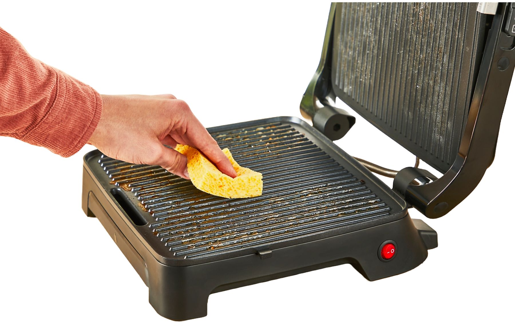 Eine Person reinigt einen schmutzigen Grill mit einem gelben Schwamm. Der Grill ist schwarz und hat einen roten Einschaltknopf.