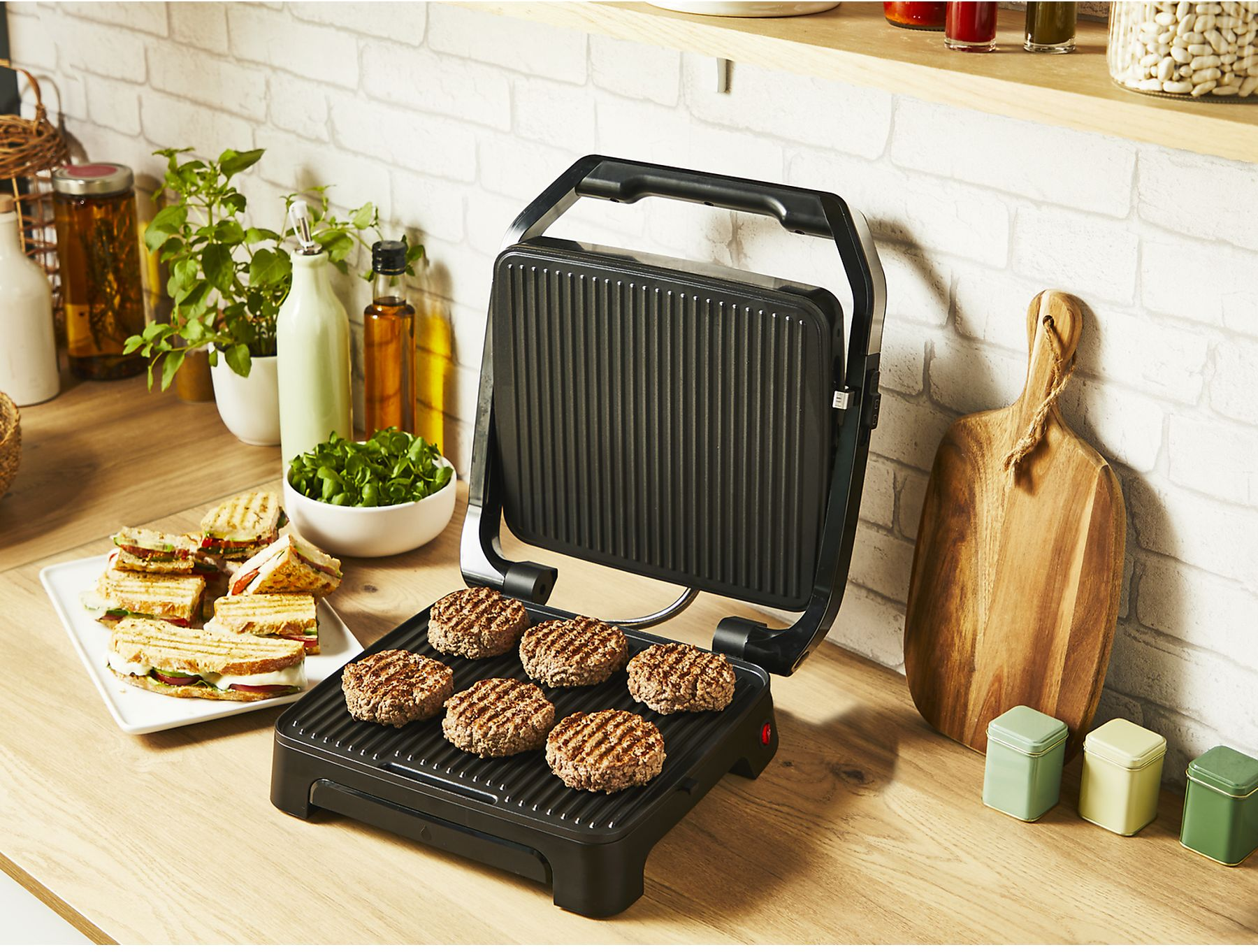 Ein schwarzer Grill mit Burgern steht auf einer Holztheke. Sandwiches und Gemüse sind daneben. Gläser, Flaschen und ein Schneidebrett.