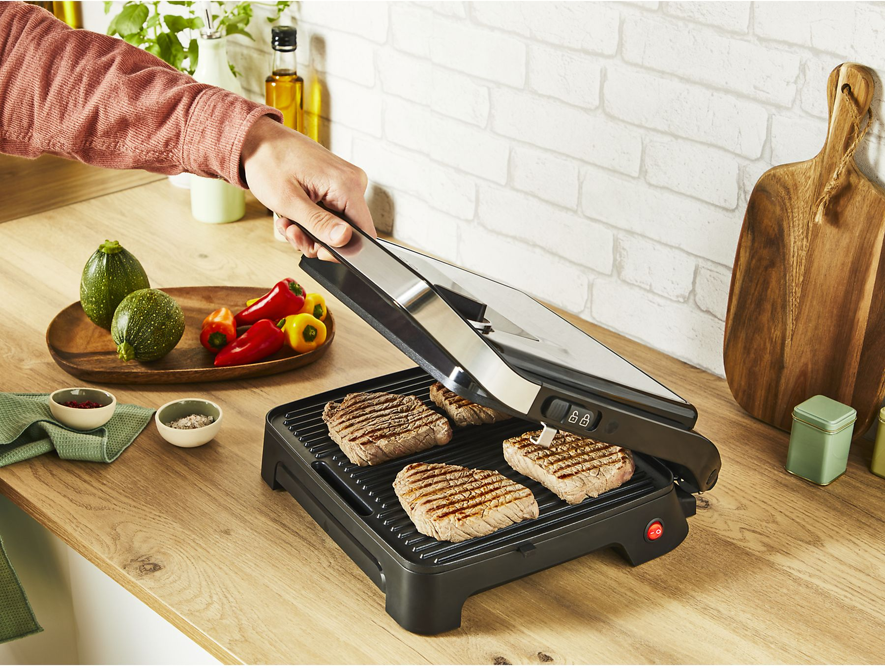 Eine Person grillt Steaks auf einem schwarzen Grill. Gemüse und Gewürze liegen auf der Holztheke. Weiße Ziegelmauer im Hintergrund.
