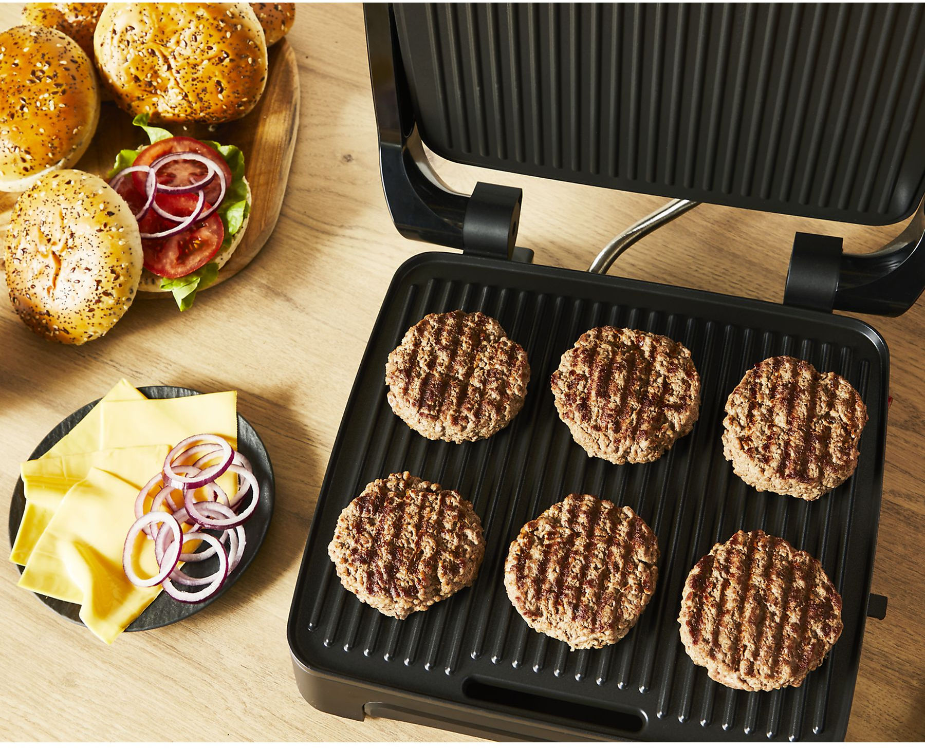 Burger-Patties auf einem Grill, mit Brötchen, Käse und Zwiebelringen. Zutaten für Burger sind auf einem Holztisch angeordnet.