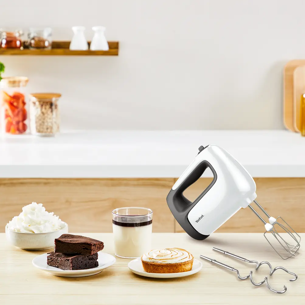 Un frullatore a mano bianco e nero con prodotti da forno su un tavolo. Brownies, una crostata e un bicchiere di dessert sono visibili.