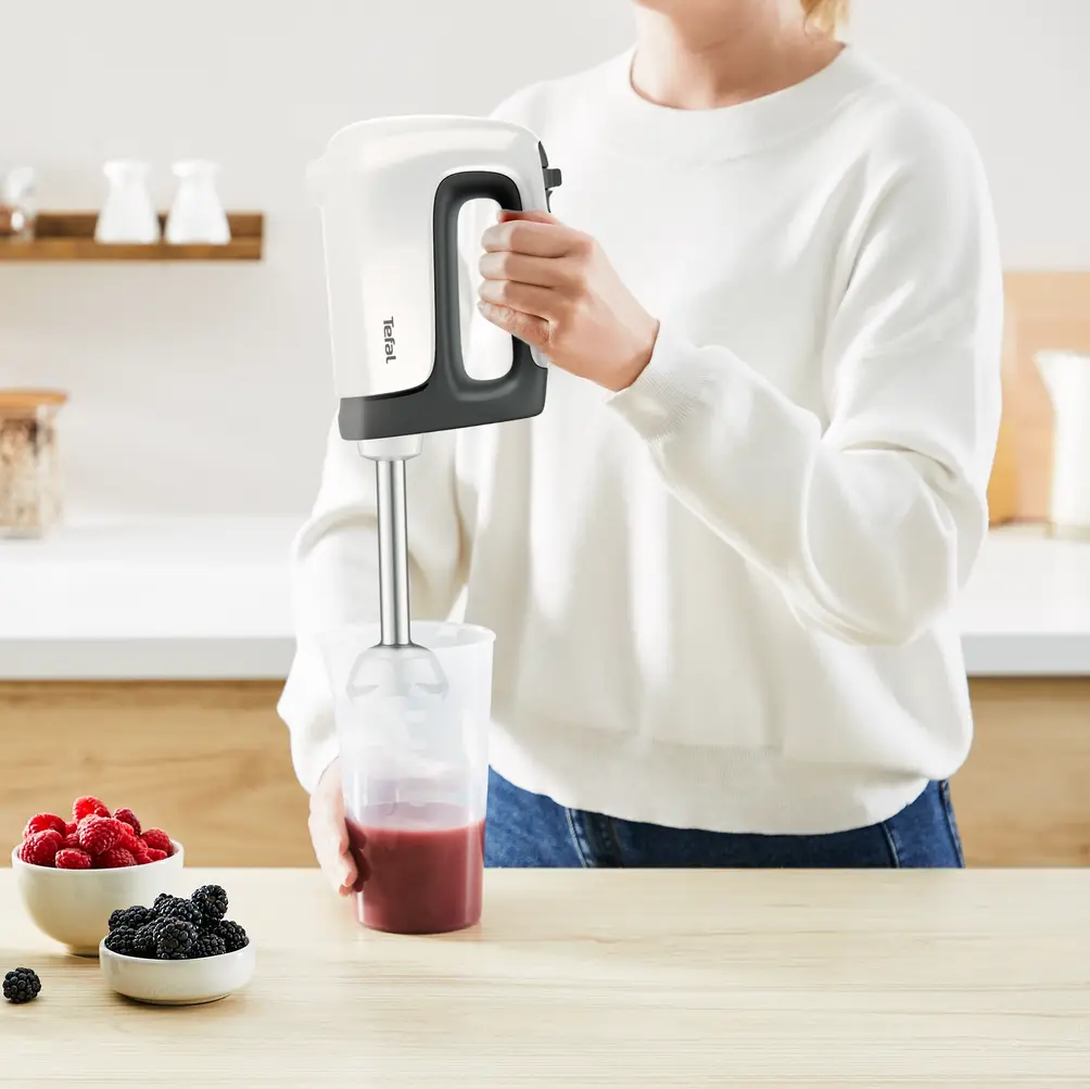 Una persona usa un frullatore a immersione Tefal. Le bacche sono in ciotole su un tavolo di legno. Il frullatore mescola liquido rosso in un bicchiere.
