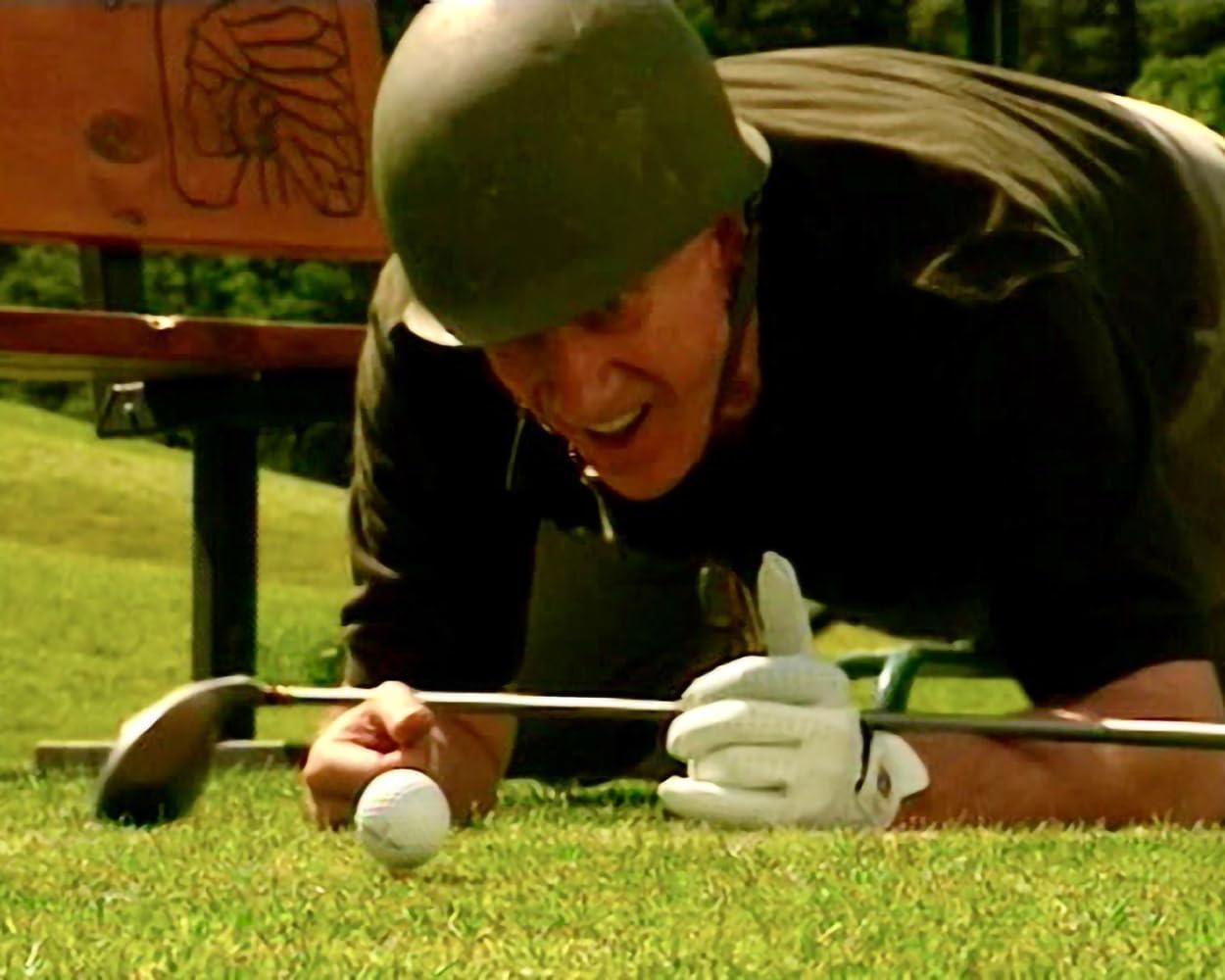 Ein Mann mit Helm kniet auf Gras und benutzt einen Golfschläger, um einen Ball zu schlagen. Er trägt einen Handschuh. Im Hintergrund ist eine Bank.