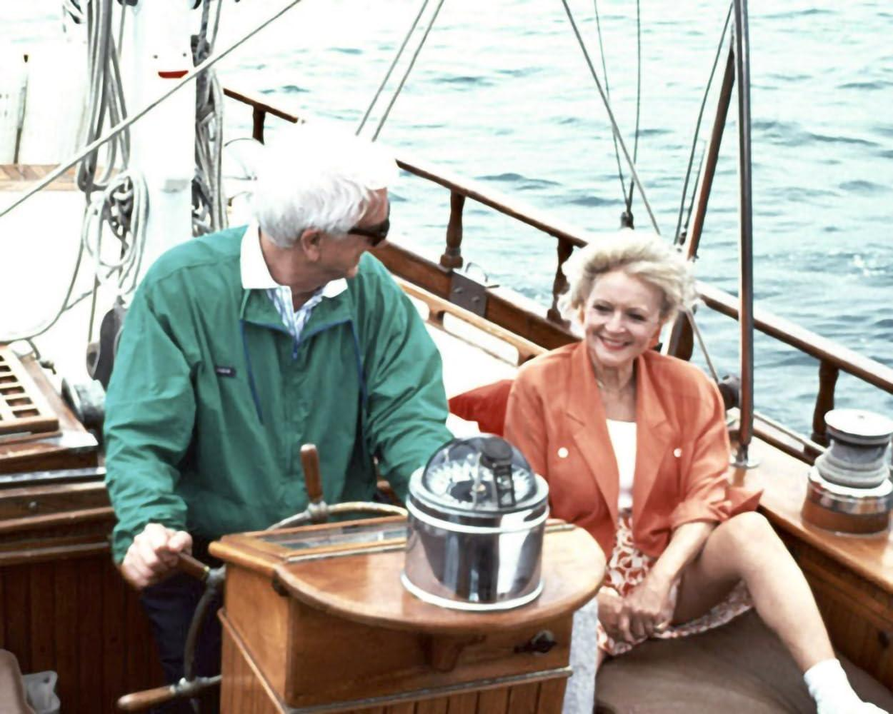 Ein Mann steuert ein Boot, eine Frau sitzt daneben. Der Mann trägt Grün, die Frau Orange. Im Hintergrund ist das Meer.