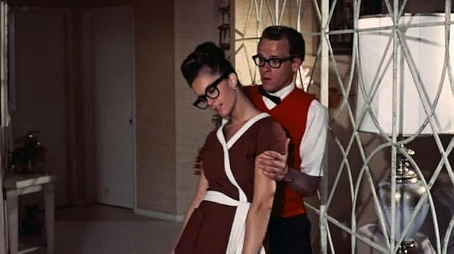 Ein Mann mit Brille und roter Weste stützt eine Frau mit Brille und braunem Kleid. Eine Lampe und eine dekorative Platte sind dahinter.