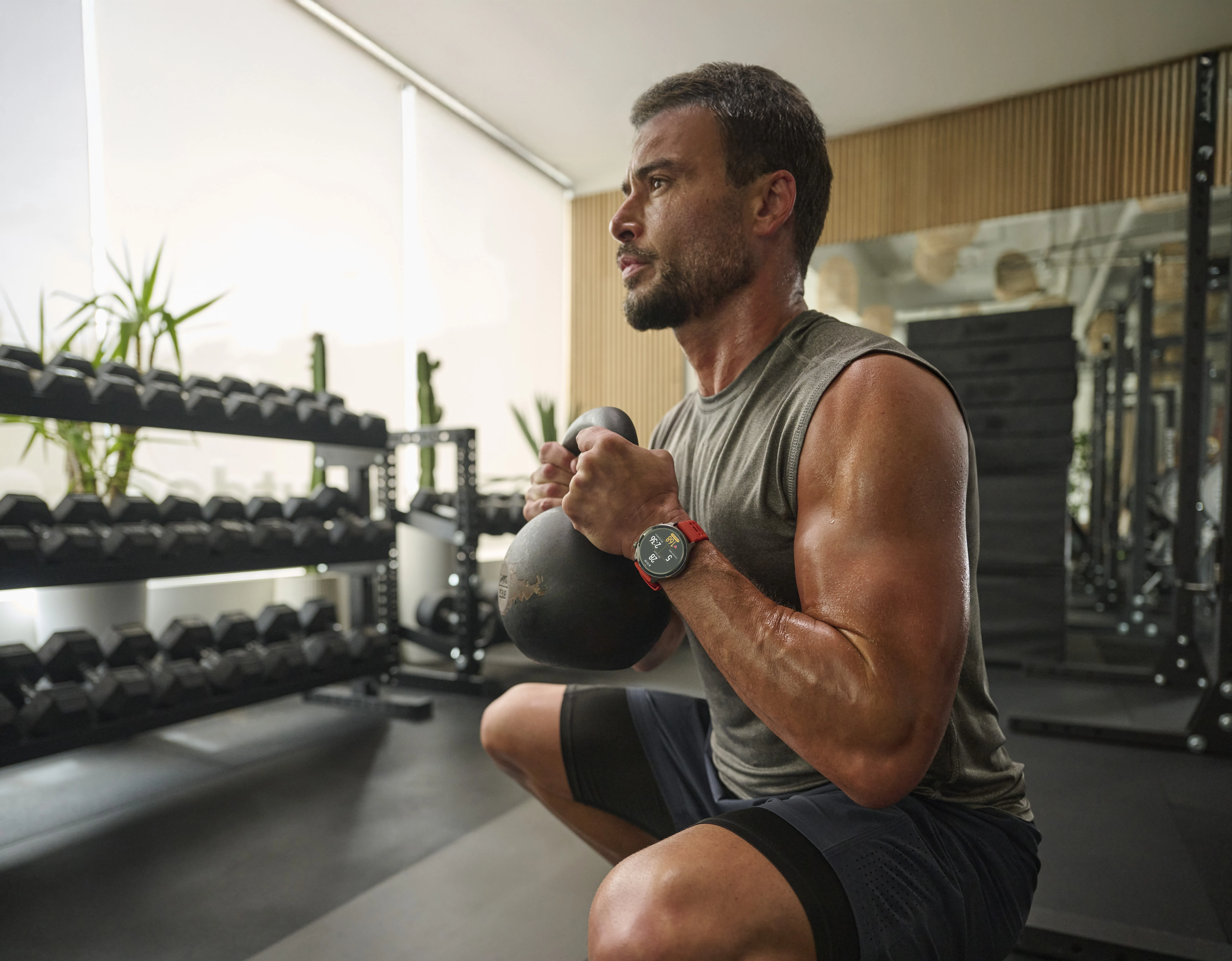 Uomo in palestra si accovaccia con kettlebell. Manubri su rack, orologio al polso, sudore visibile.