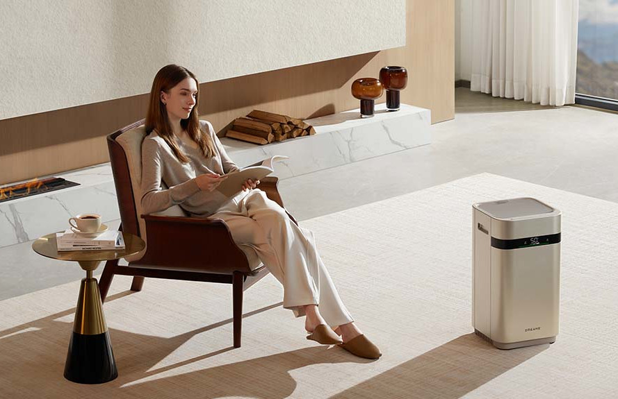 Kobieta czyta, siedząc w fotelu. Stolik kawowy, oczyszczacz powietrza i kominek w pokoju.