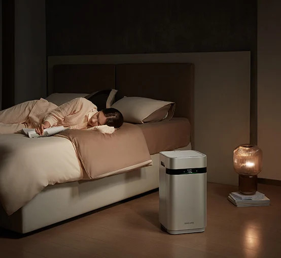Osoba śpi na łóżku. Oczyszczacz powietrza stoi w pobliżu. Lampa pali się na stoliku nocnym. Pokój jest słabo oświetlony.