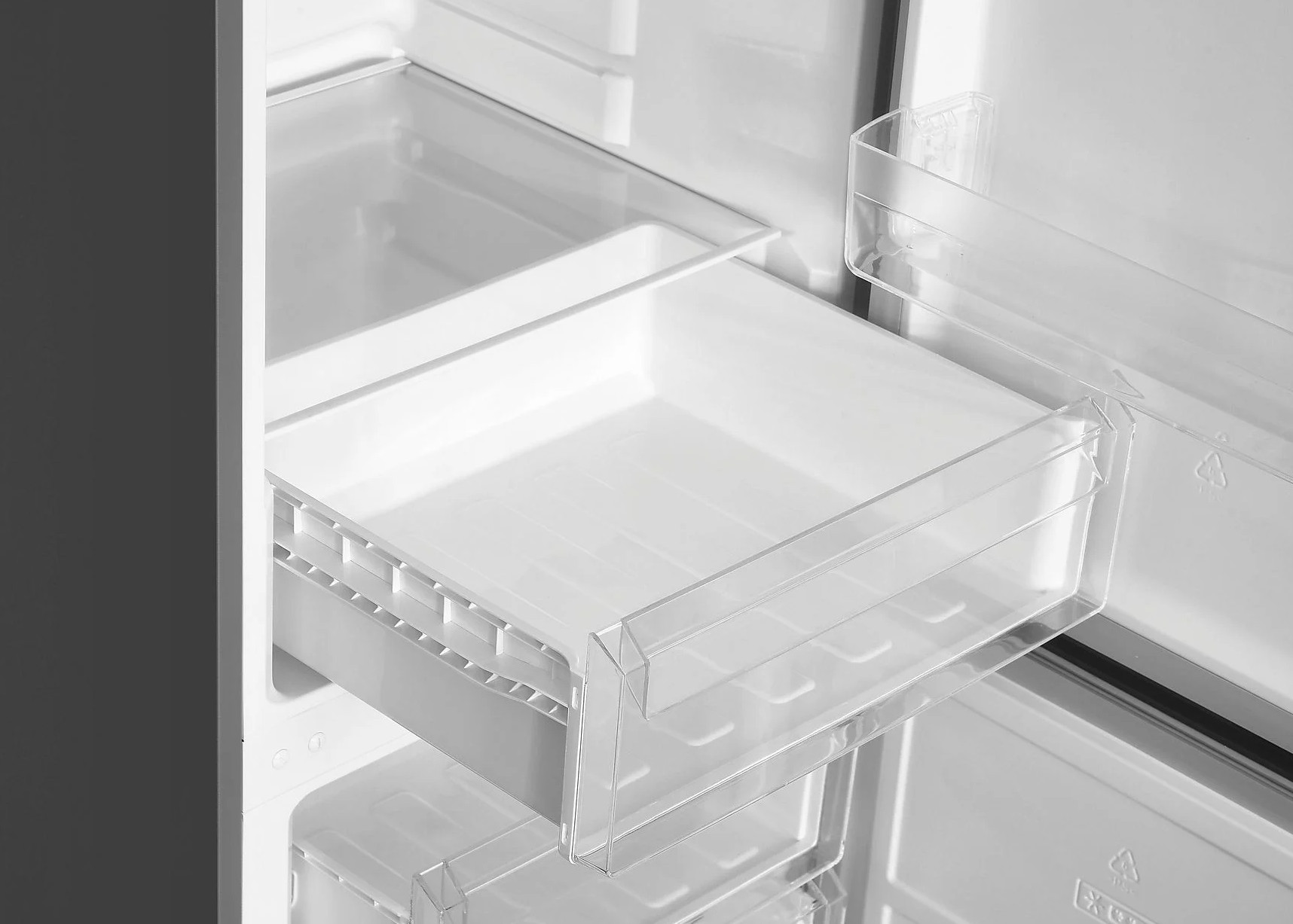 Wewnątrz białej lodówki wysunięta jest przezroczysta szuflada. Widoczne są inne białe przegrody.