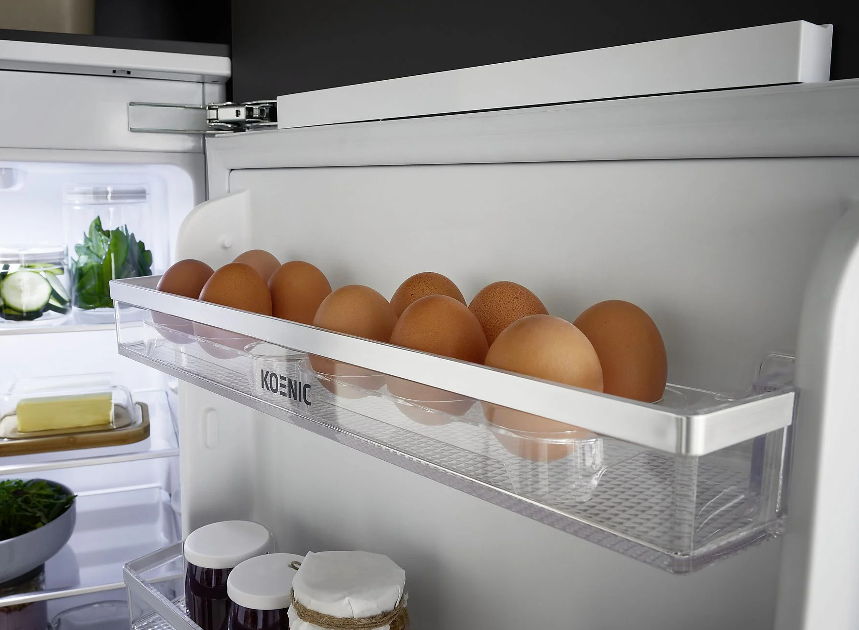 Otwarta lodówka z brązowymi jajkami na półce, widoczne są również warzywa i masło. Marka Koenic jest na półce.