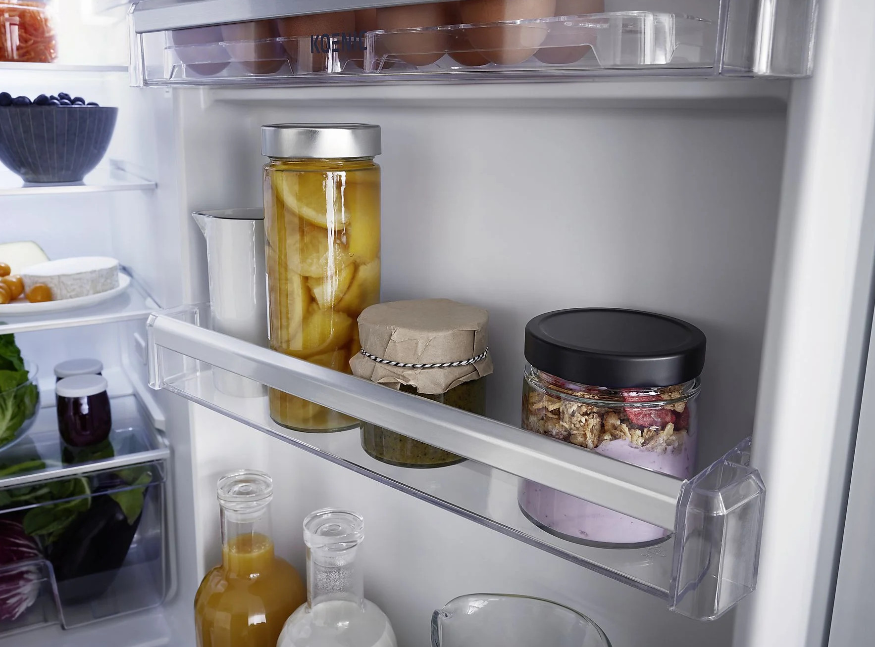 W lodówce: słoiki z jedzeniem, jajka, miska z jagodami, ser i butelki z sokiem. Przezroczyste półki.