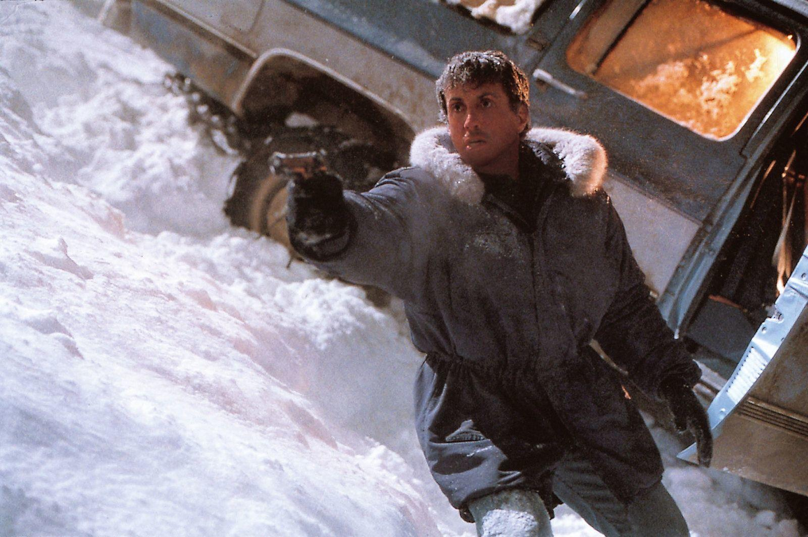 Ein Mann in einer Pelzjacke hält eine Waffe im Schnee, nahe einem beschädigten Fahrzeug.