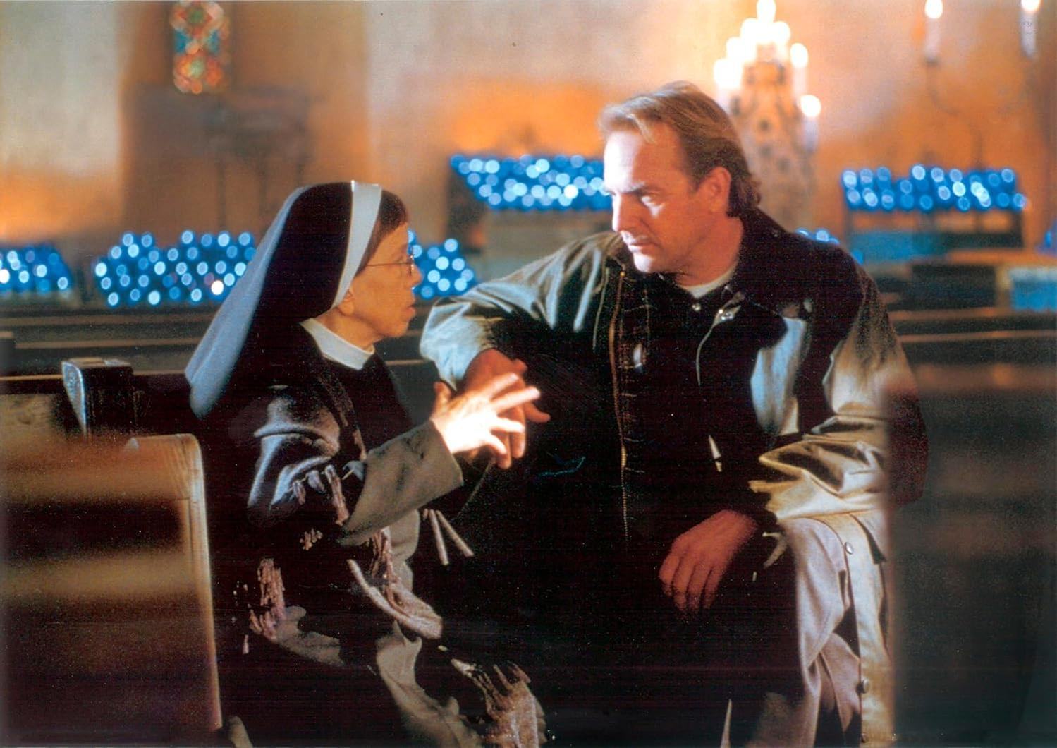 Eine Nonne und ein Mann sitzen in einer schwach beleuchteten Kirche auf einer Kirchenbank und unterhalten sich.