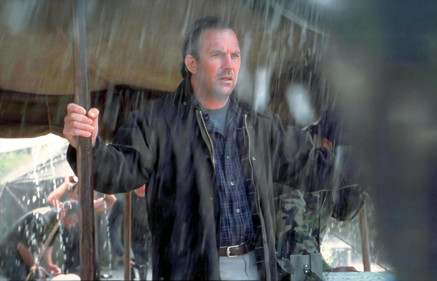 Mann in dunkler Jacke und kariertem Hemd steht im starken Regen und hält Stange. Andere im Hintergrund.